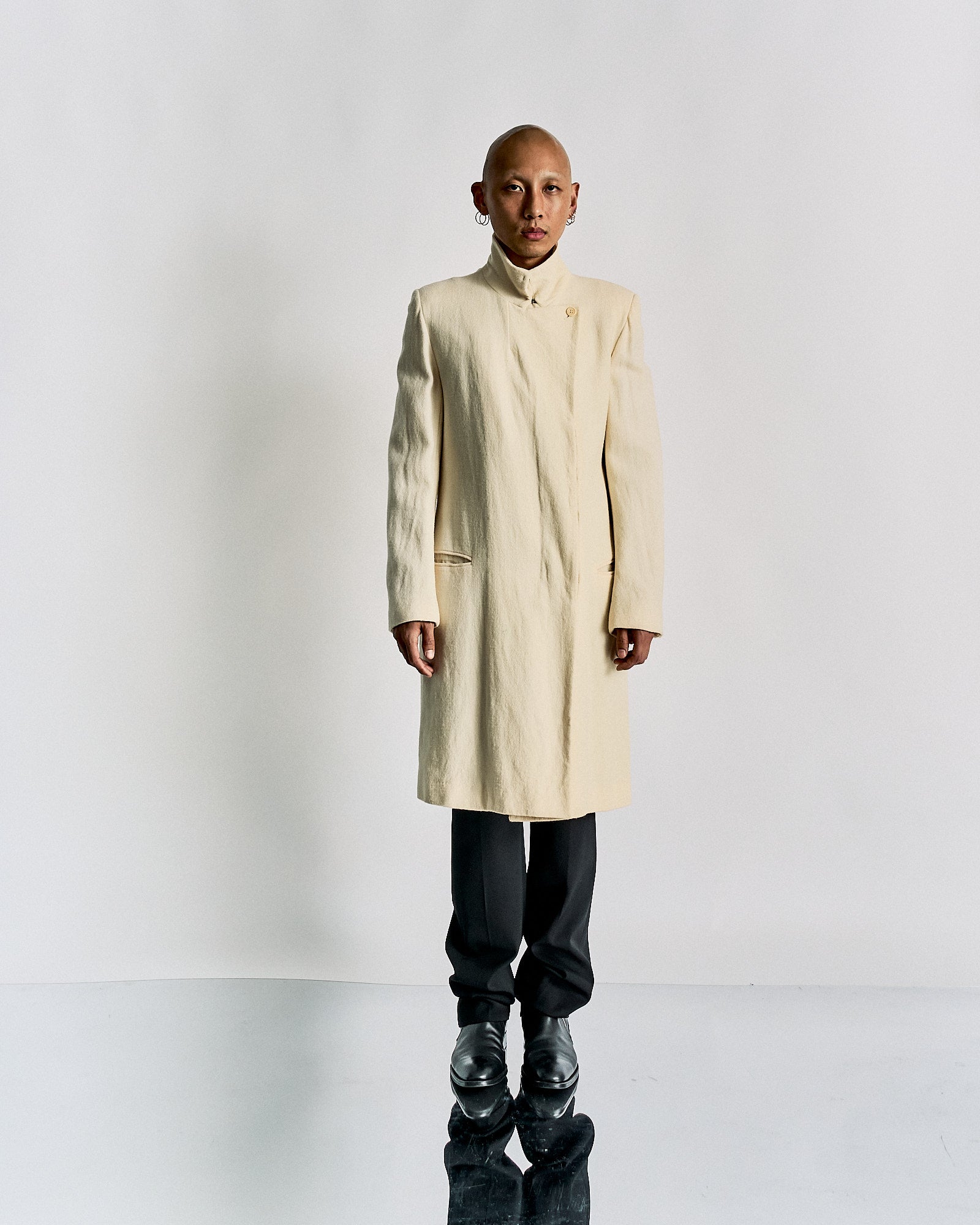 Person wearing a long cream-coloured Ann Demeulemeester coat and black pants stands against a plain white background. 