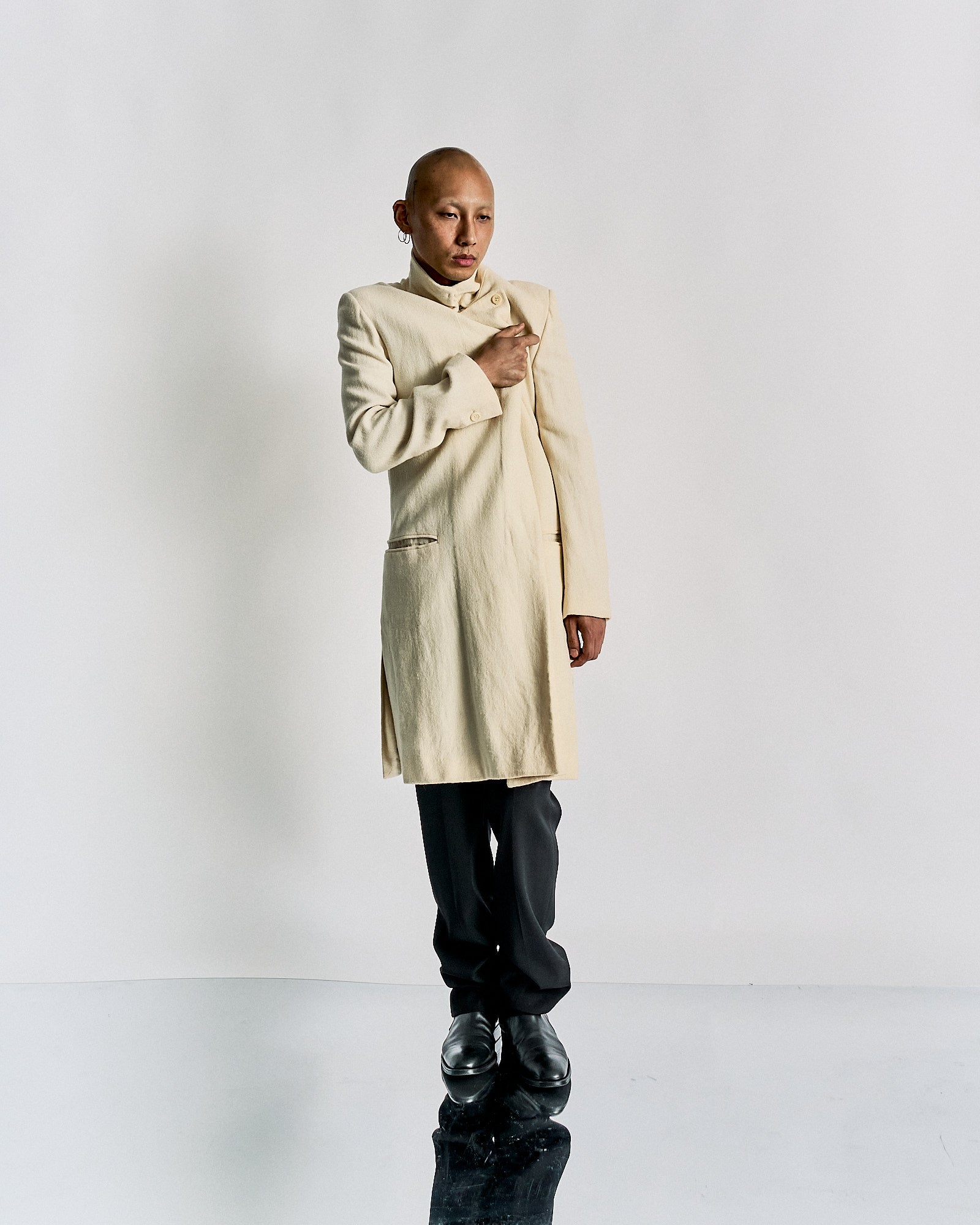 A person stands confidently against a plain background, wearing a cream-coloured Ann Demeulemeester coat over black pants and shoes. They project a poised, modern elegance.
