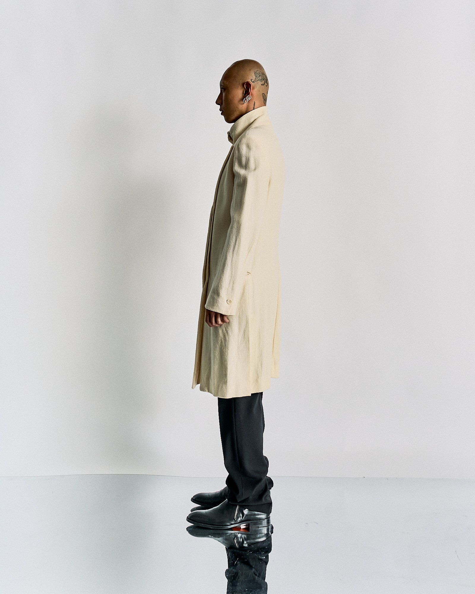 Side profile of a person wearing a long beige Ann Demeulemeester coat, black pants, and shiny black shoes, standing on a reflective surface against a plain background.
