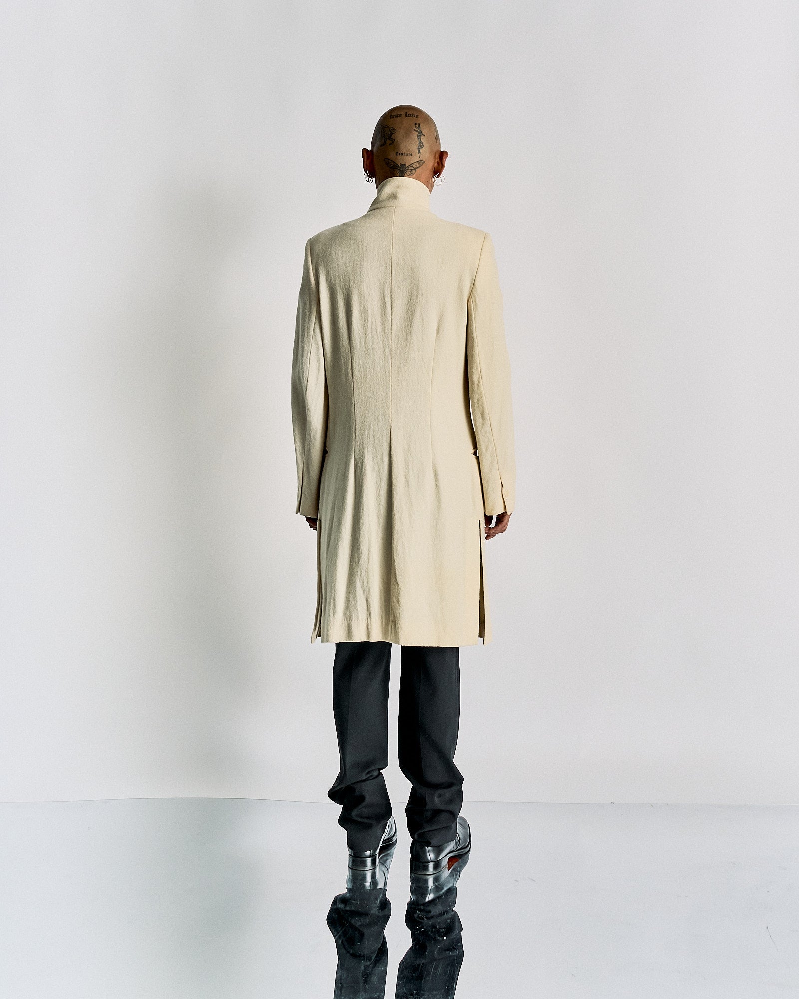 Man with tattooed head and unique hairstyle wearing a long, cream-coloured Ann Demeulemeester coat with black pants and shiny boots, standing on a reflective surface.