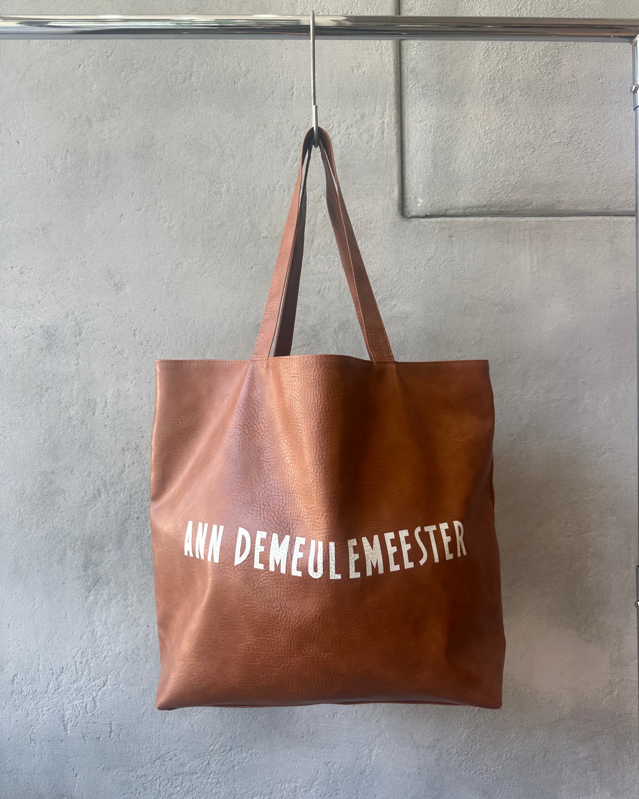 Brown tote bag with 'ANNE DEMEULEMEESTER' text hanging on a gray wall.
