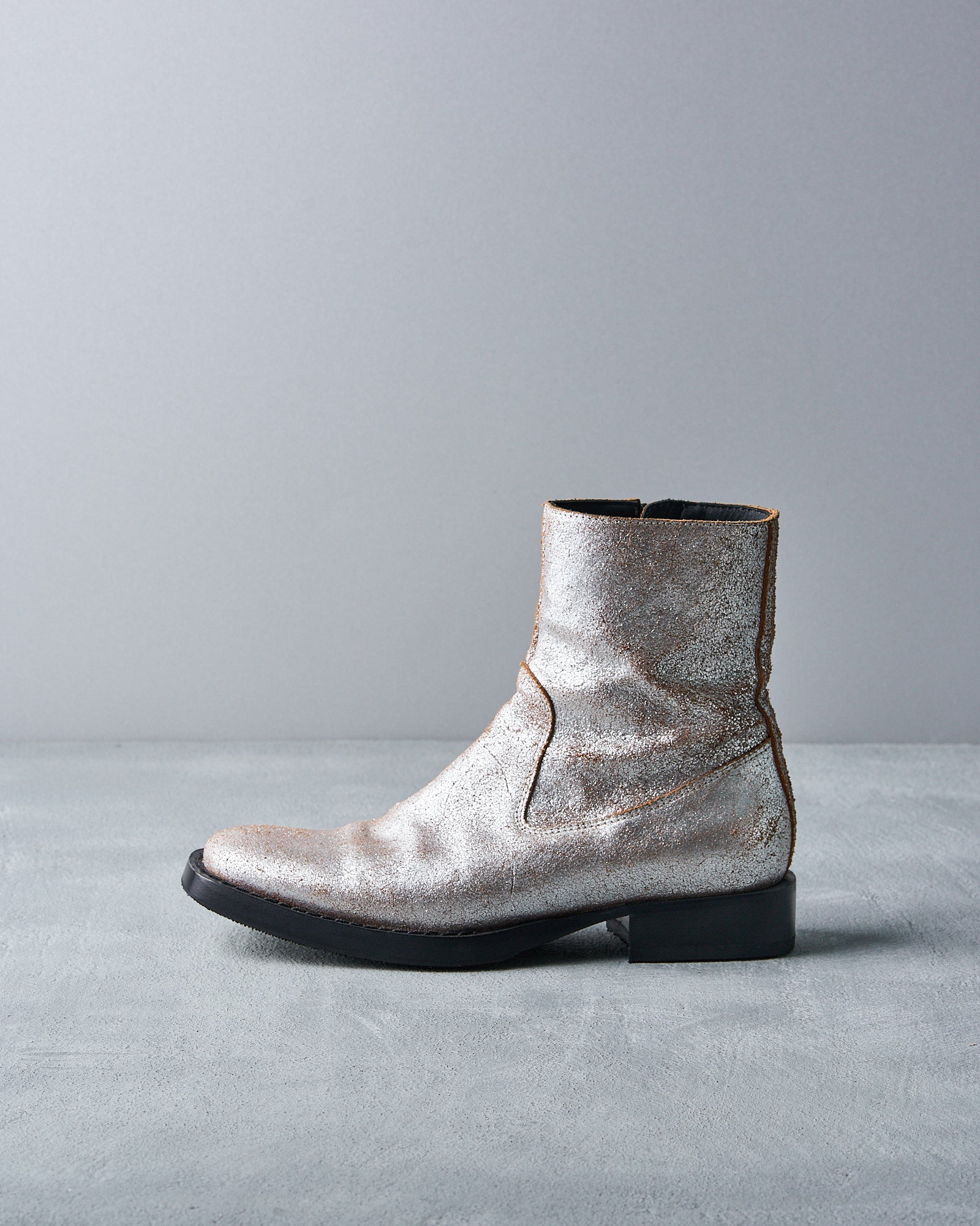 Silver metallic Ann Demeulemeester ankle boot with a textured finish on a gray surface and backdrop. The boot has a black sole, creating a sleek, modern look.