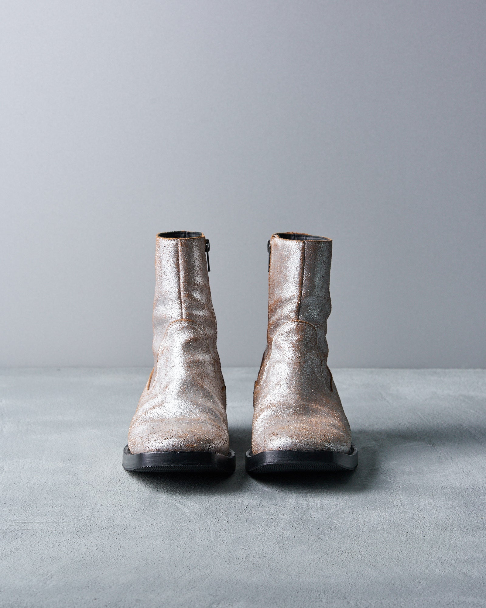 Silver ankle boots with a metallic sheen, side zippers, and black soles, resting on a textured gray surface, conveying a stylish and modern vibe.
