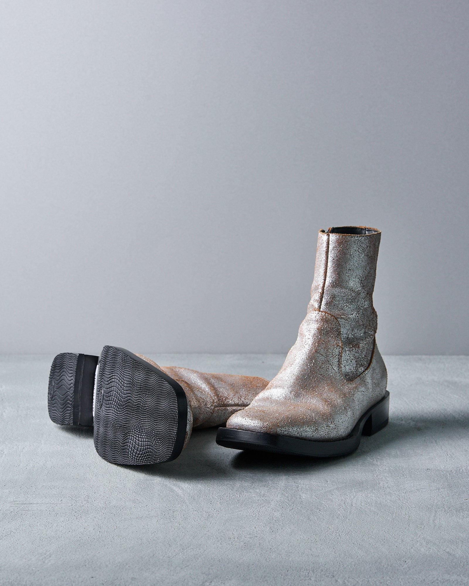 Silver, glittery Ann Demeulemeester ankle boots with black soles are displayed on a gray surface. One boot stands upright while the other lies on its side, showcasing the sole.