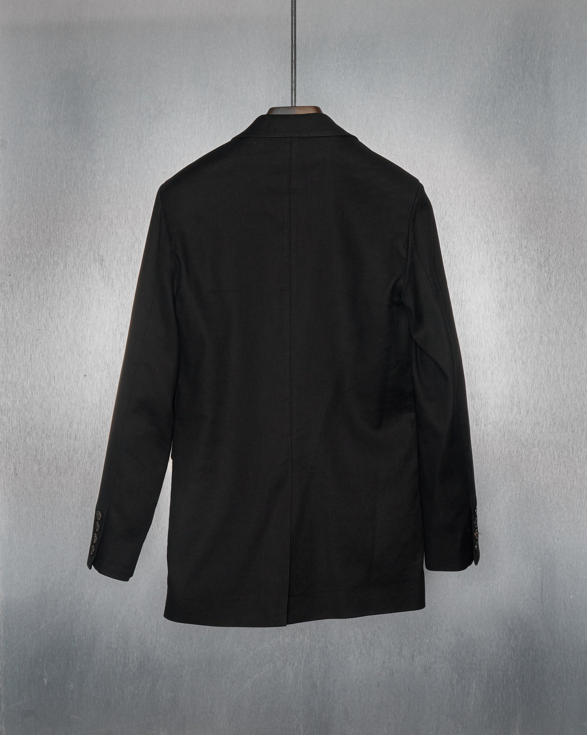 Alt text: "Back view of a black Ann Demeulemeester suit jacket hanging on a metal hanger against a light gray textured background. The jacket has a classic design."