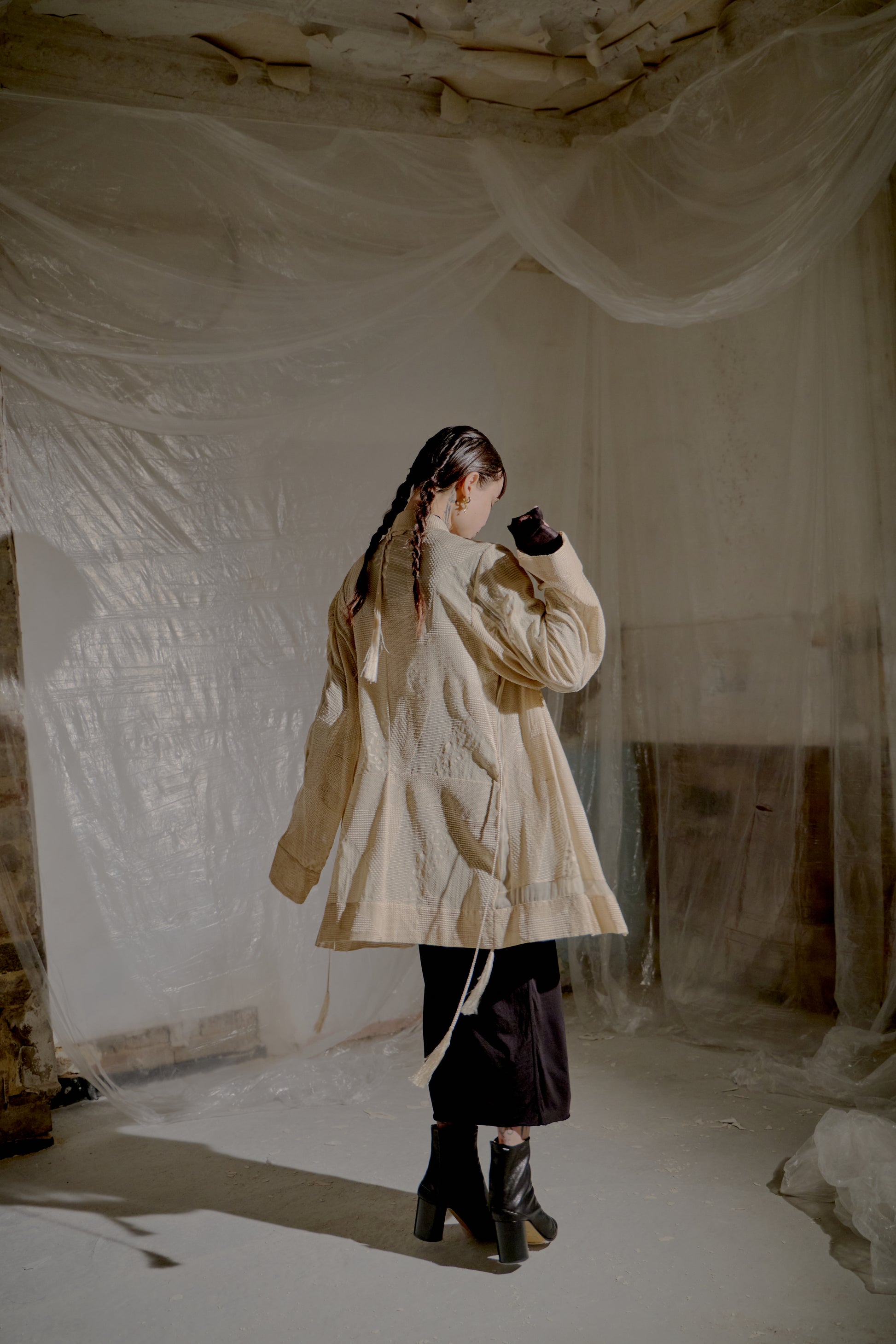 Person wearing a beige Ann Demeulemeester coat standing in a room with draped fabric