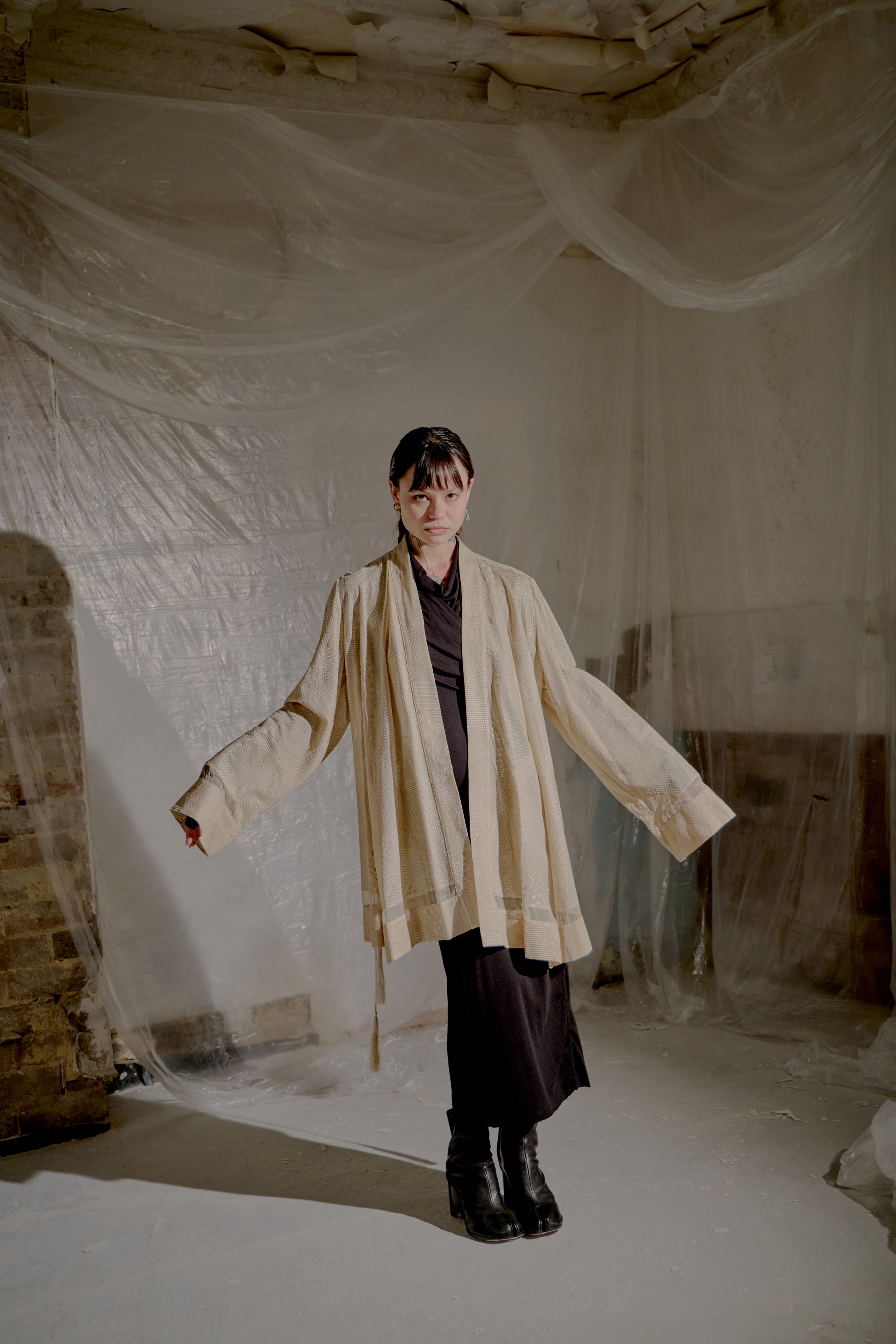 Person wearing a beige Ann Demeulemeester coat in a room with white plastic sheeting on the walls