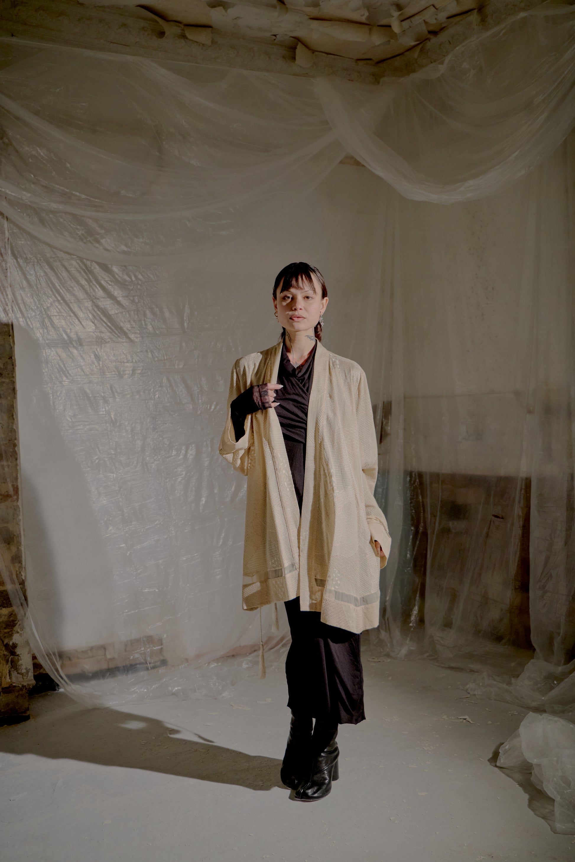 Person wearing a beige Ann Demeulemeester coat in a minimalistic indoor setting with white fabric.