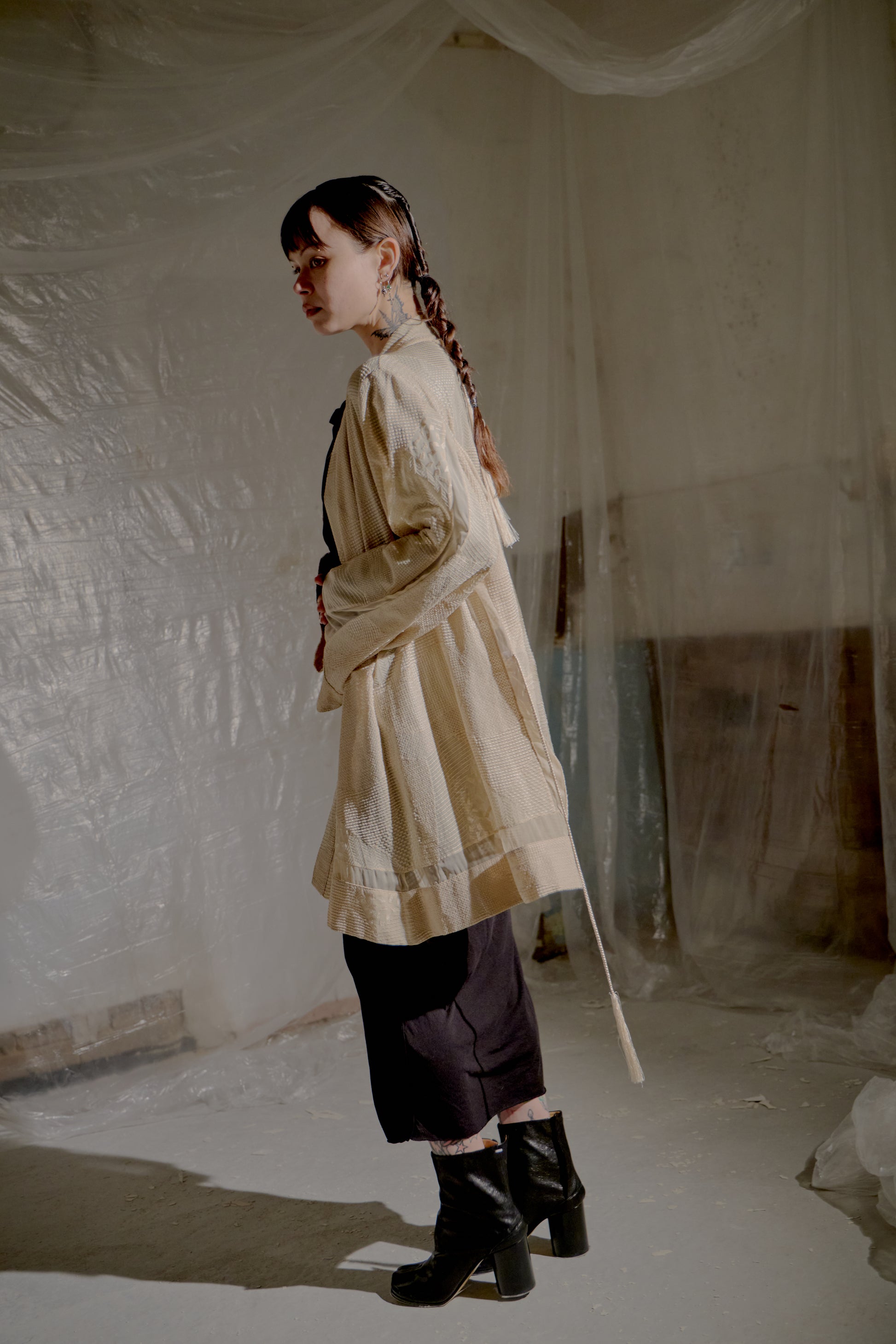 Person wearing a beige Ann Demeulemeester coat and dark pants in a minimalistic setting