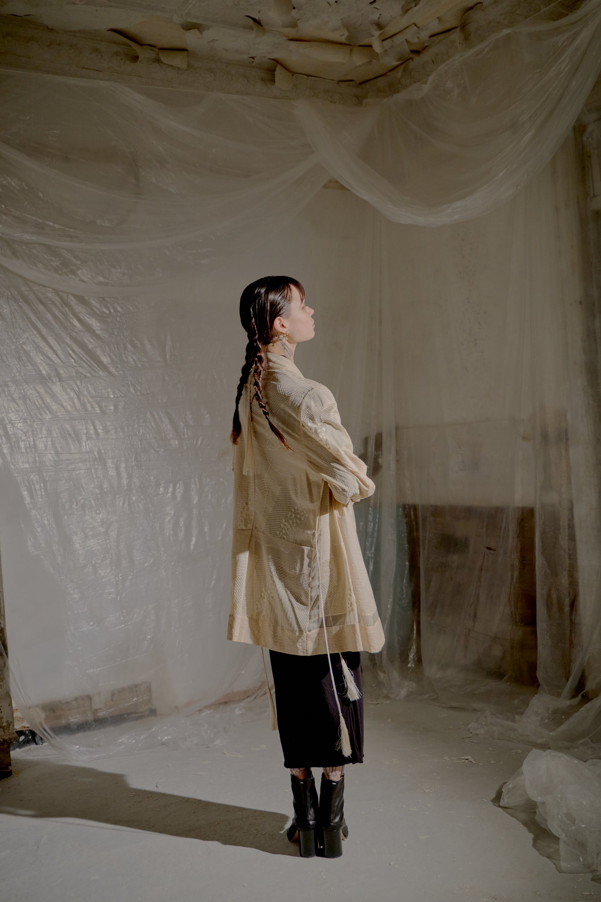 Person standing in a room wearing a light Ann Demeulemeester coat and in a room with draped fabric walls and ceiling