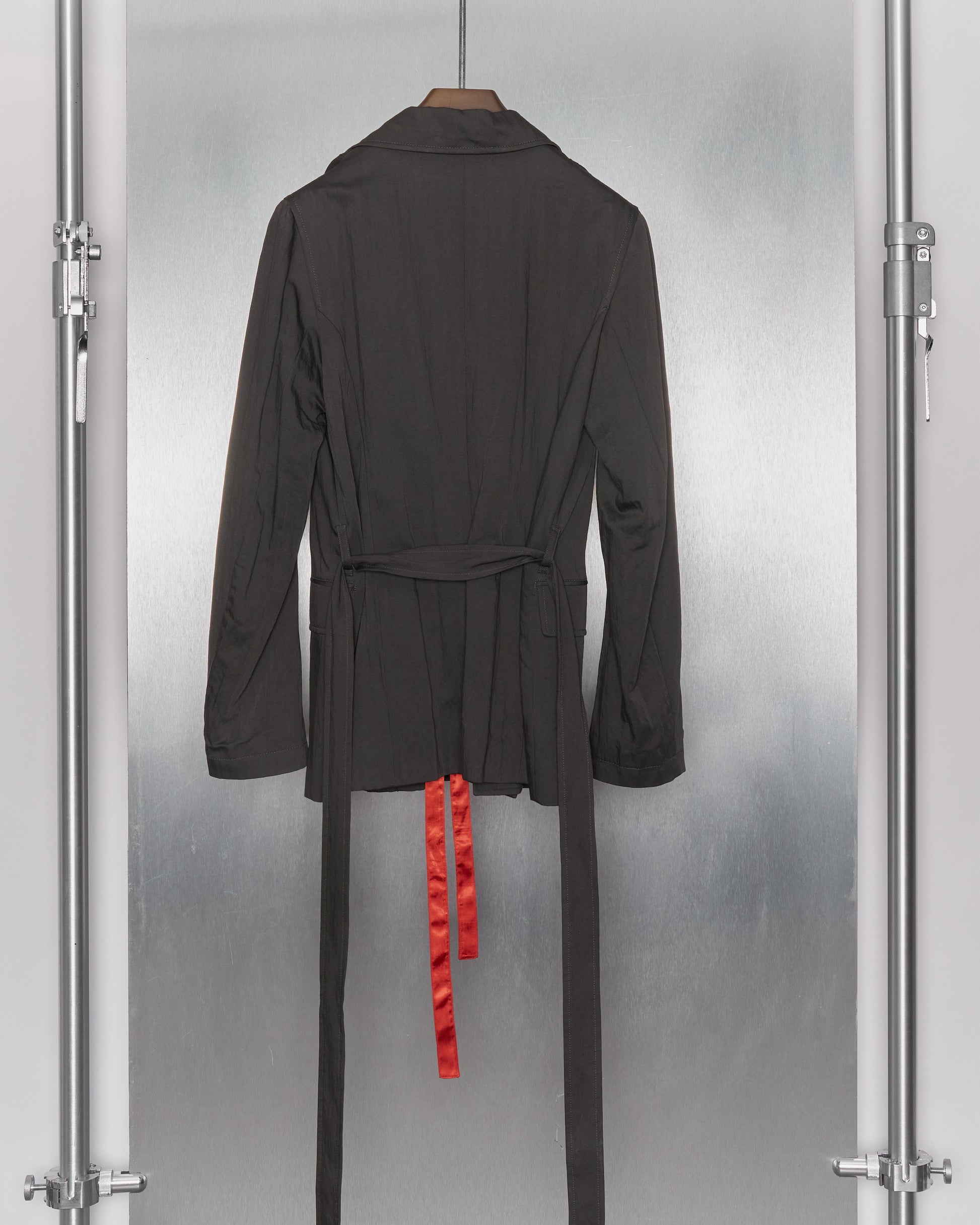 A grey Ann Demeulemeester hanging on a hook, with a red satin ribbon protruding from the bottom. The background is a textured gray surface, creating a dramatic contrast.