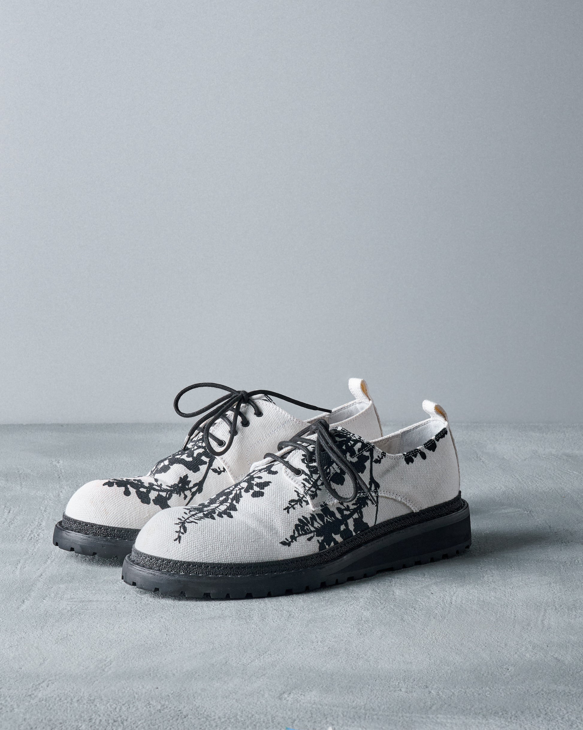 White Ann Demeulemeester shoe with black floral patterns and black laces sits on a gray surface against a light gray background, conveying a stylish and modern feel.