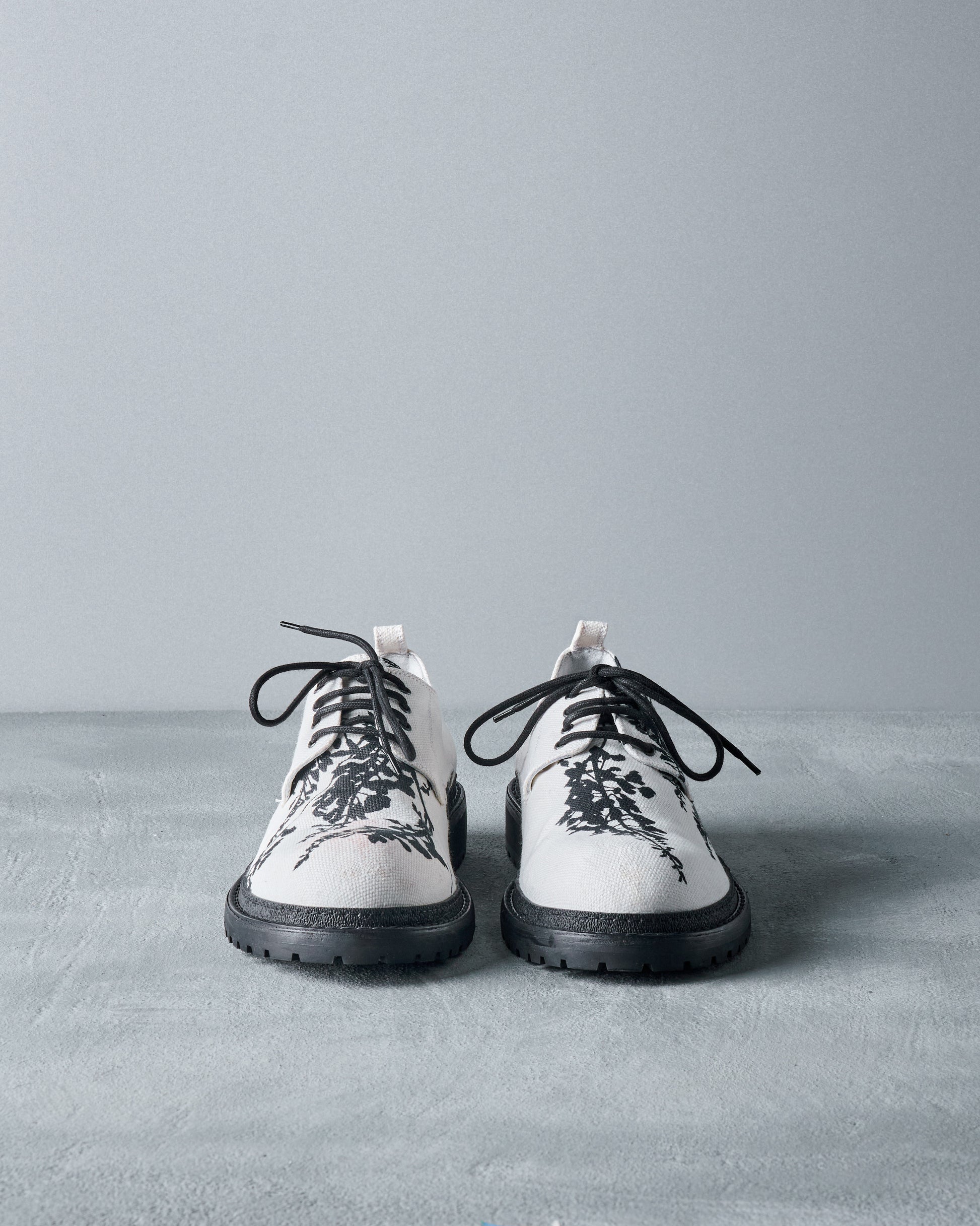 White lace-up Ann Demeulemeester shoes with black soles and floral patterns on the toes. They are set against a gray textured background, conveying a minimalist, artistic vibe.