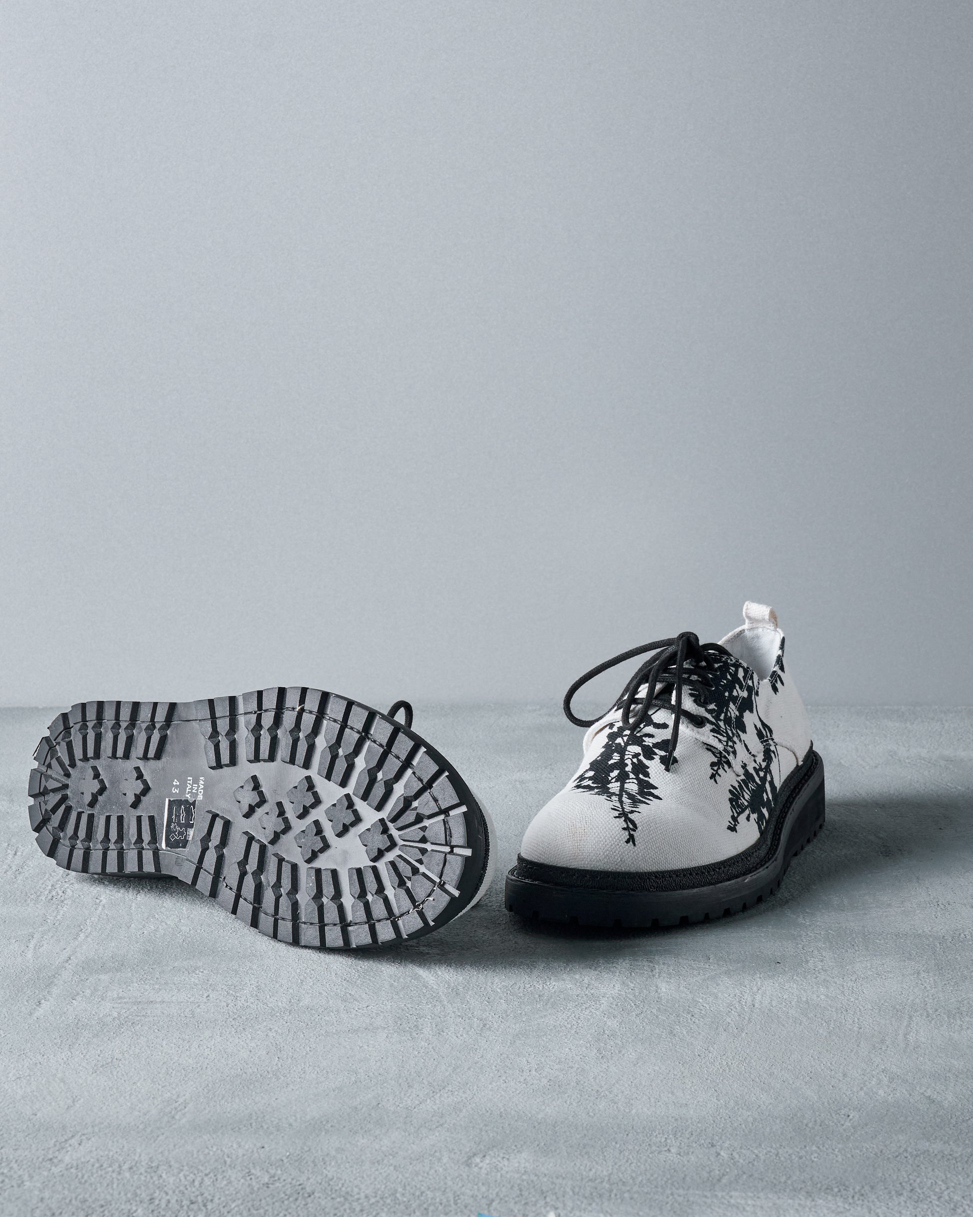 A pair of stylish white Ann Demeulemeester shoes, one upright and one showing the thick, rugged sole. Decorated with black fern patterns, conveying a modern, artistic tone.