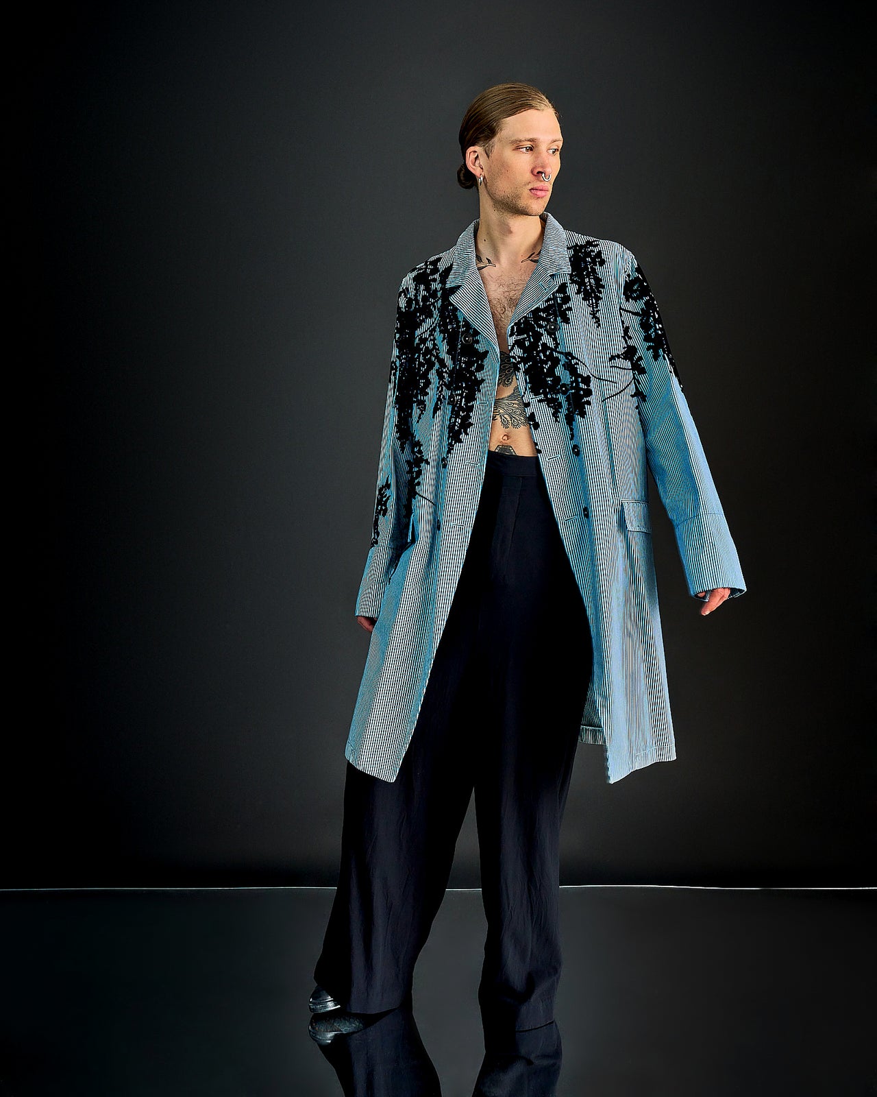 A male model walks down the runway in a light blue Ann Demeulemeester trench coat with black floral patterns, layered over a striped shirt and black pants. He wears a black beret.