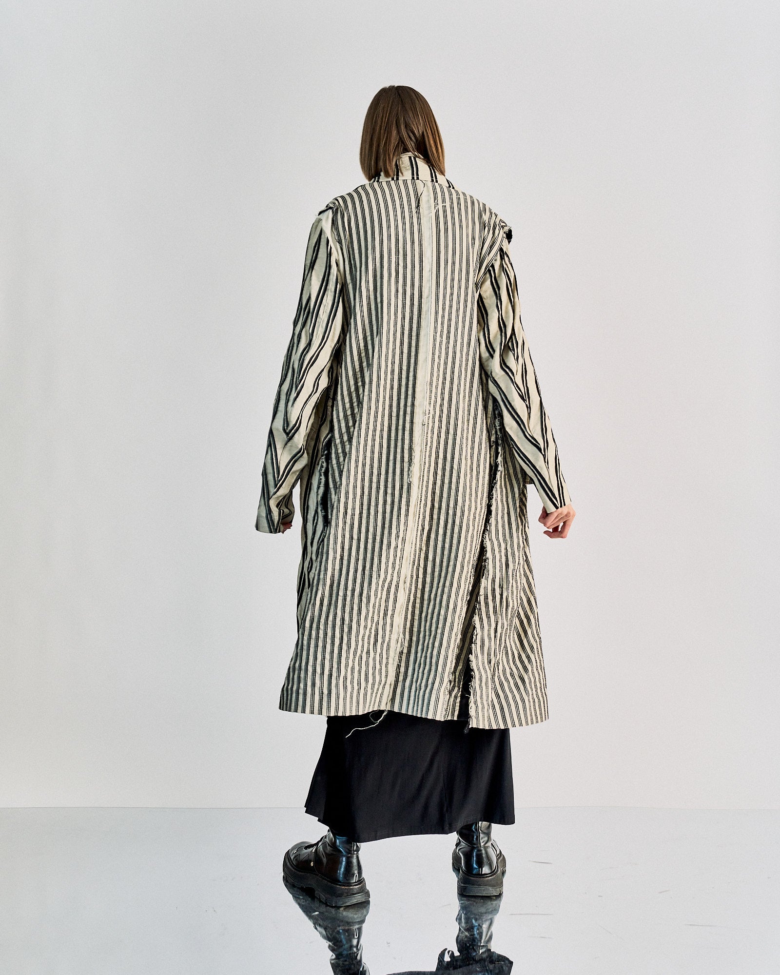 Person in a black skirt and oversized striped Ann Demeulemeester coat, seen from the back. They wear chunky black shoes, standing on a reflective surface.