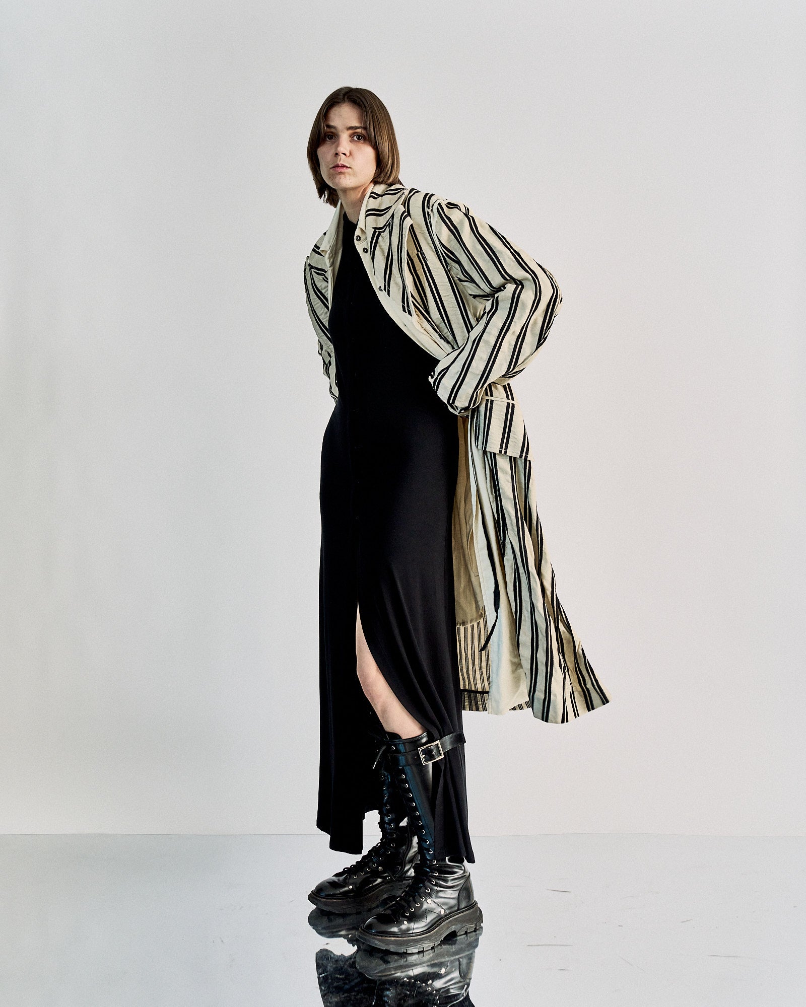 A person in a long black dress, striped Ann Demeulemeester coat, and chunky black boots stands on a reflective surface.