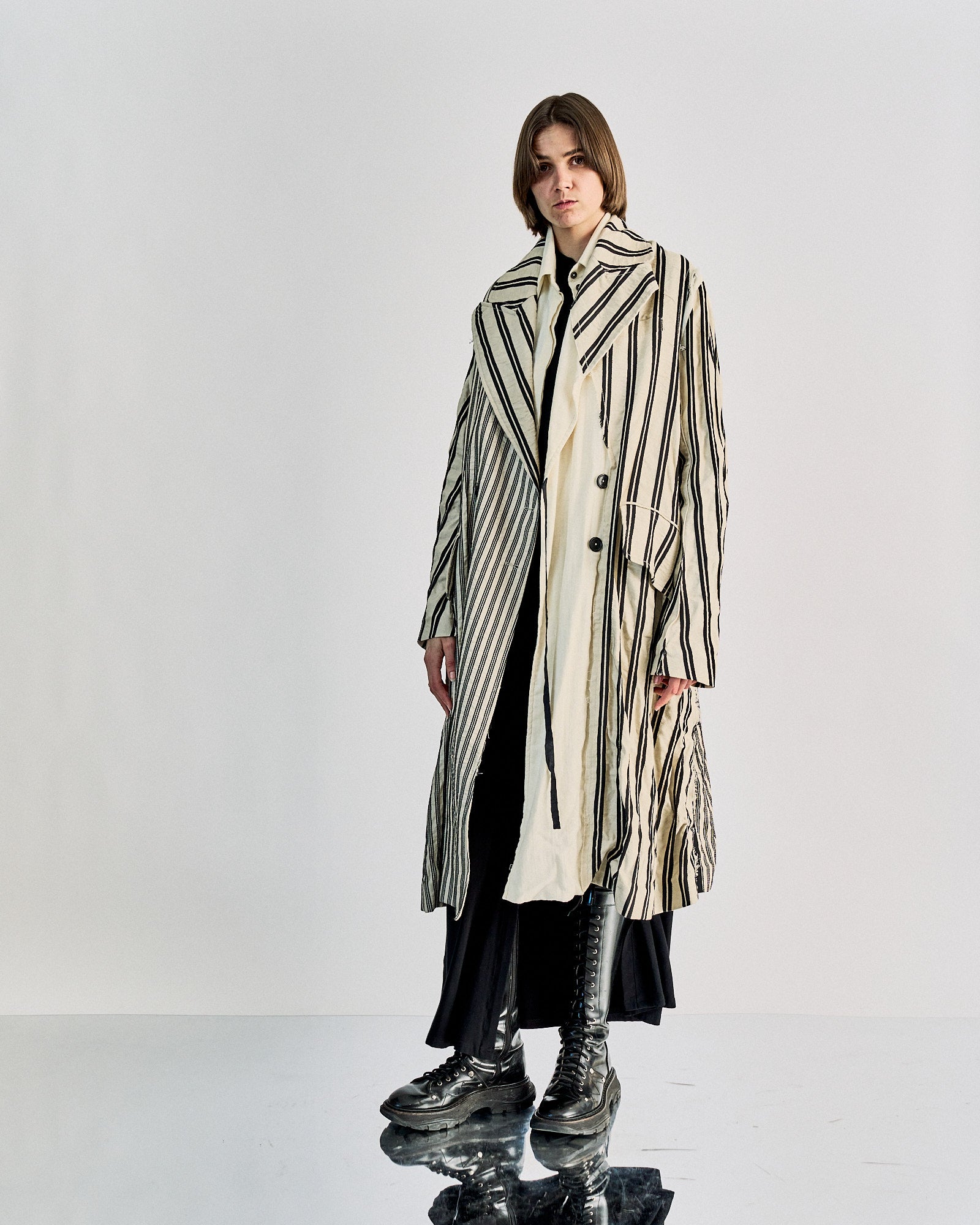 Person wearing a long, cream-coloured Ann Demeulemeester coat with vertical black stripes, paired with a black skirt and tall lace-up boots, standing on a reflective floor.