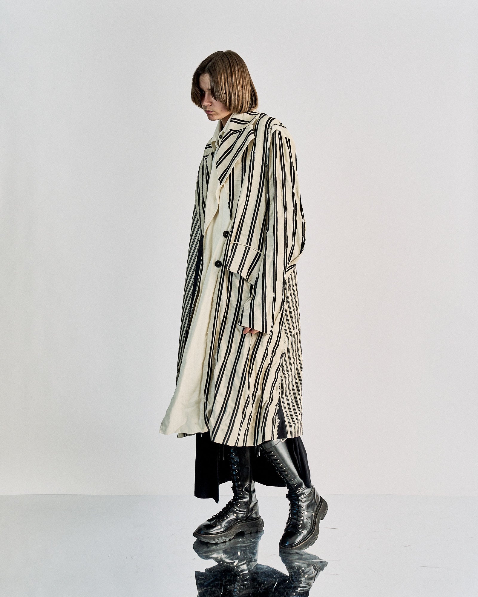 A model wearing a striped two-piece Ann Demeulemeester coat with a wide peak lapel, the outer piece is striped while the inner jacket is solid ecru. The coat is styled over the shoulders with a relaxed fit.