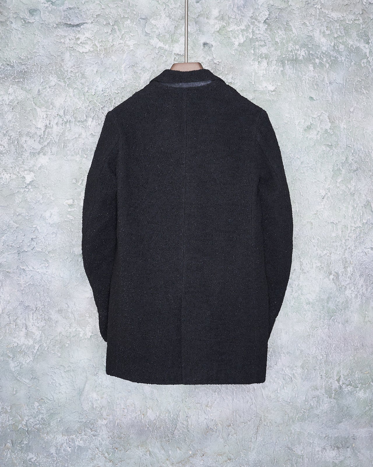 A black double-breasted Ann Demeulemeester coat with buttons is neatly hung on a textured gray wall. The coat exudes a formal and classic style.
