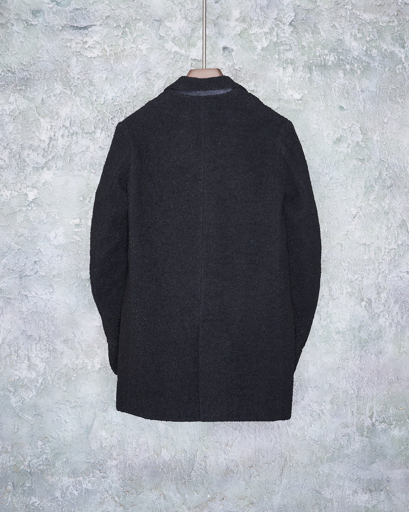 Black woollen Ann Demeulemeester blazer hanging against textured light gray wall, viewed from the back. The fabric appears thick, conveying a formal and elegant tone.