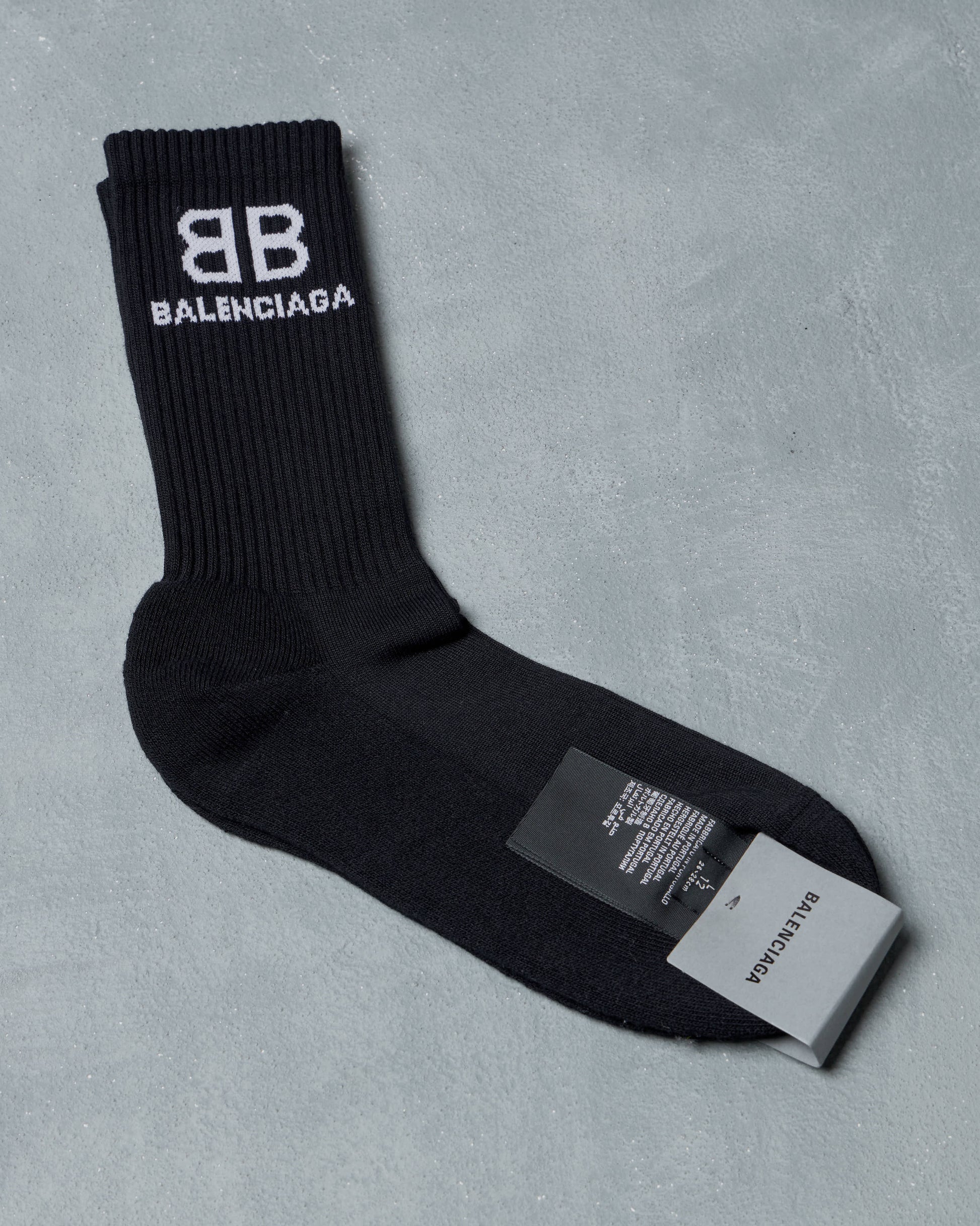 Black Balenciaga crew socks on a gray surface with "BB Balenciaga" logo in white at the top. A tag with brand details is attached at the heel. 