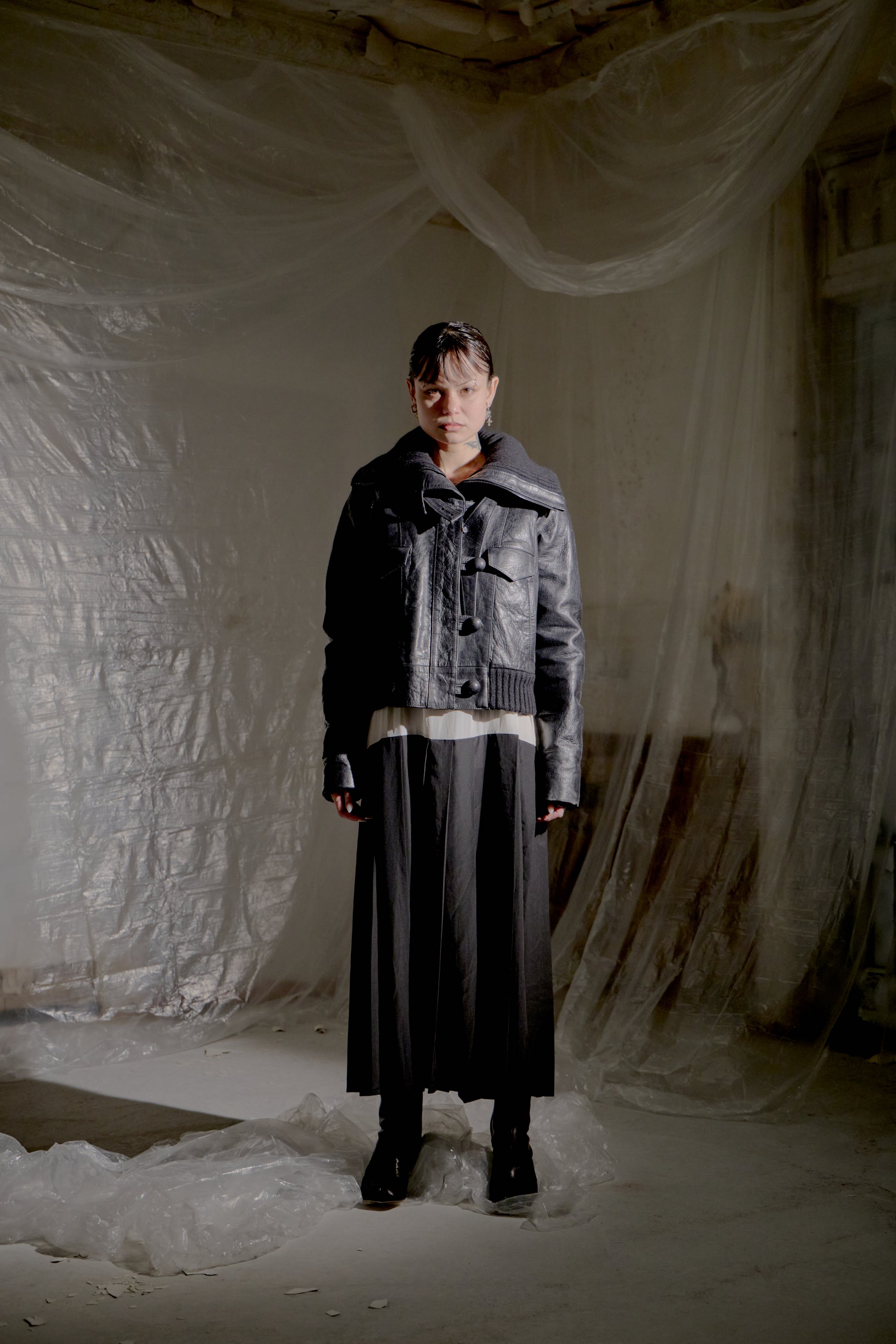 Person wearing a black leather Balenciaga jacket and skirt standing in a minimalistic setting with white draped fabric.