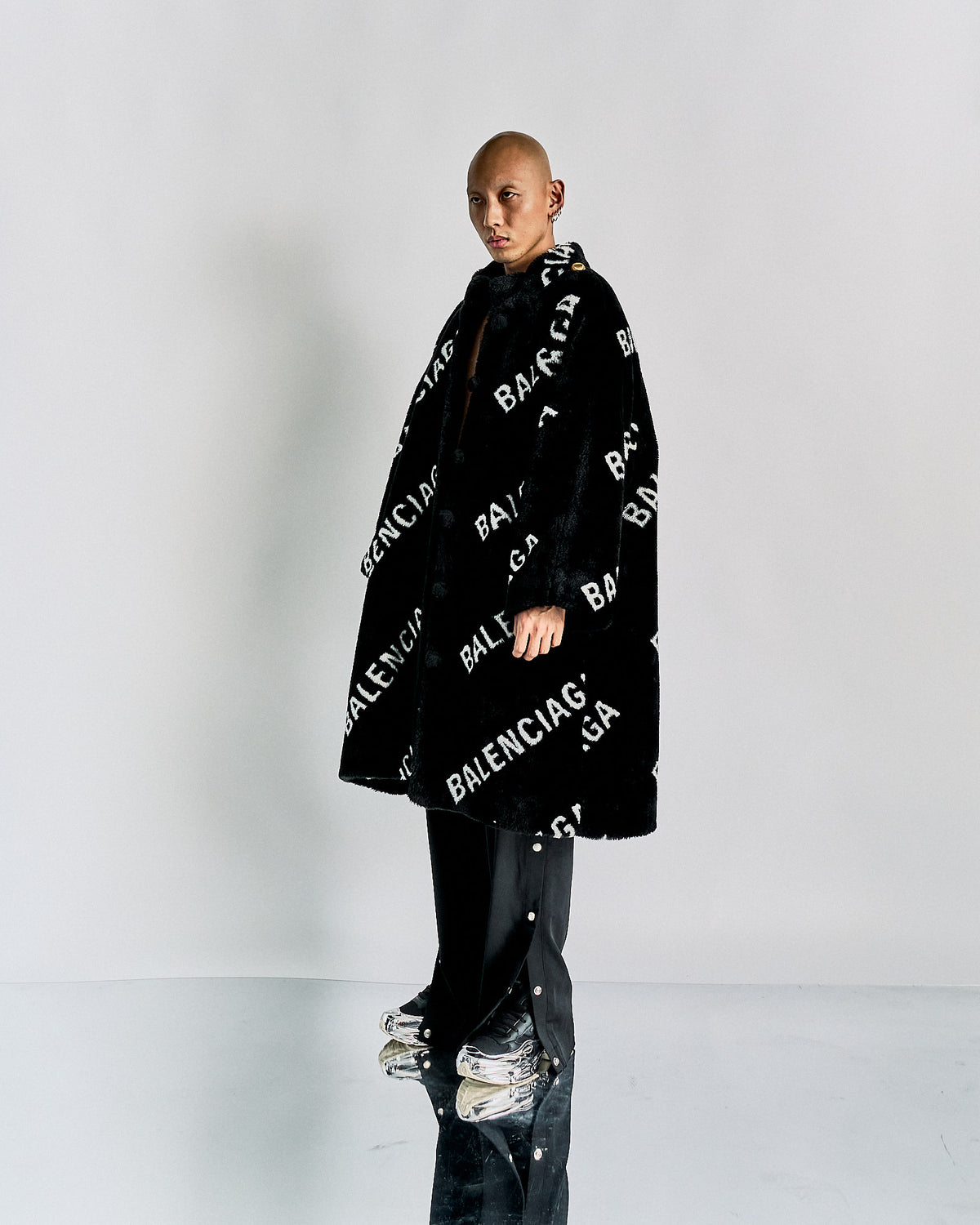A model wearing an oversized, black faux fur Balenciaga logo coat with a boxy silhouette, featuring button-front closures and internal quilted lining.
