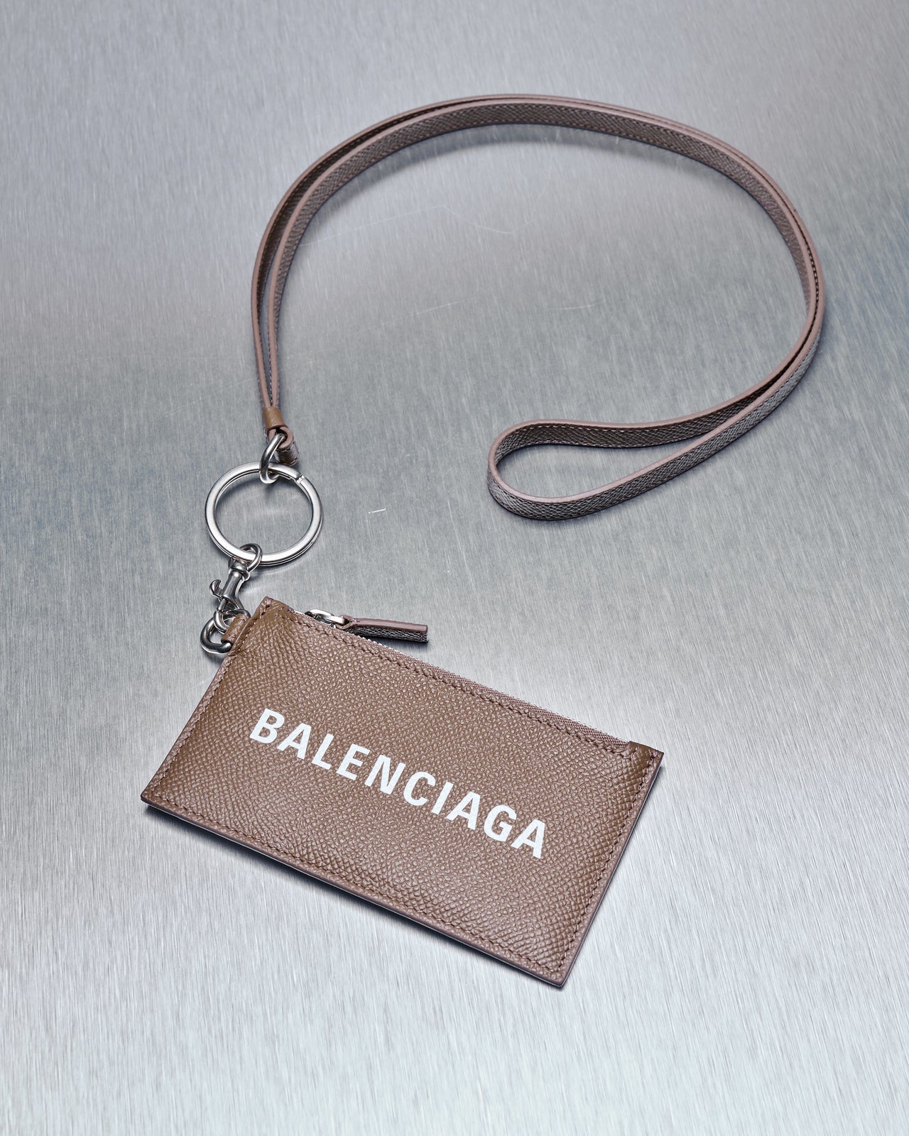 A brown grained leather card holder with a Balenciaga logo on the front, featuring a zip coin purse, four card slots, and a clip lanyard with a keychain.