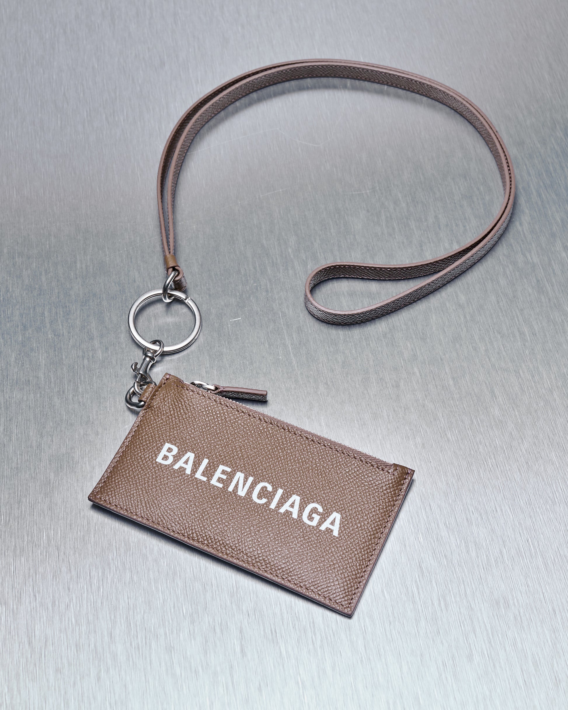 A brown grained leather card holder with a Balenciaga logo on the front, featuring a zip coin purse, four card slots, and a clip lanyard with a keychain.