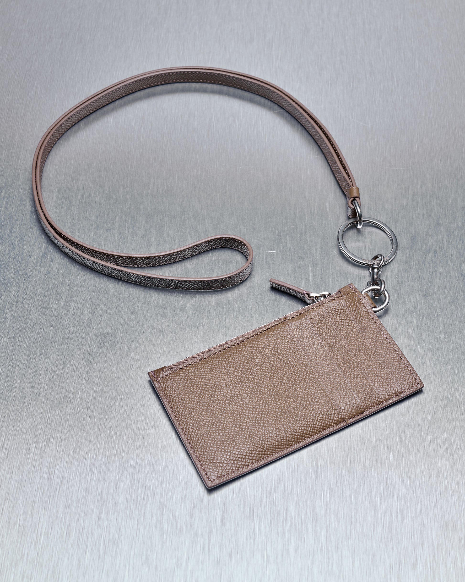 A brown leather Balenciaga lanyard with a key ring and attached cardholder lies on a smooth, metallic surface. The cardholder has a textured finish, conveying elegance.