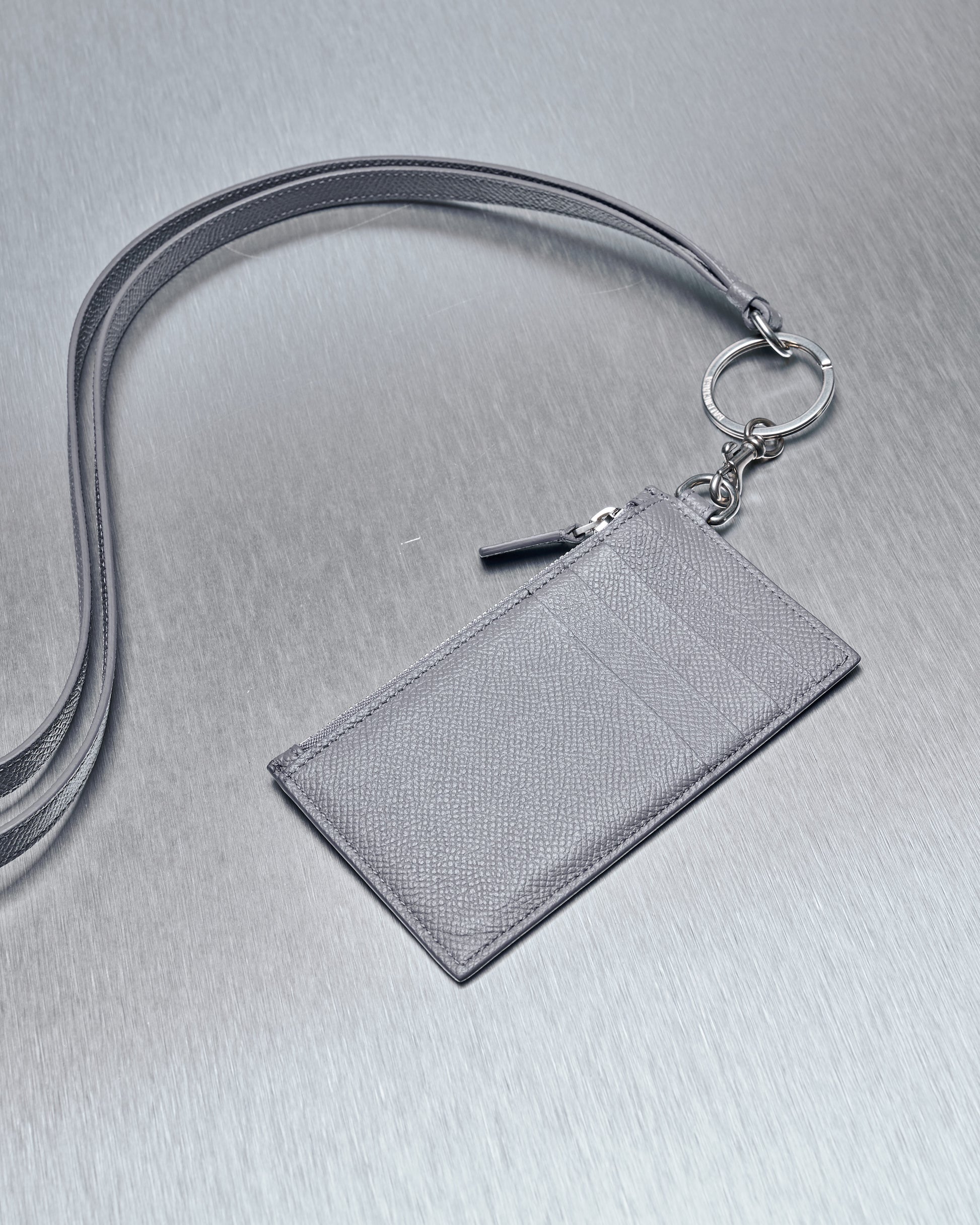 A sleek grey leather Balenciaga ID holder with a lanyard, featuring a key ring attachment. It lies flat on a metallic grey background, conveying elegance.