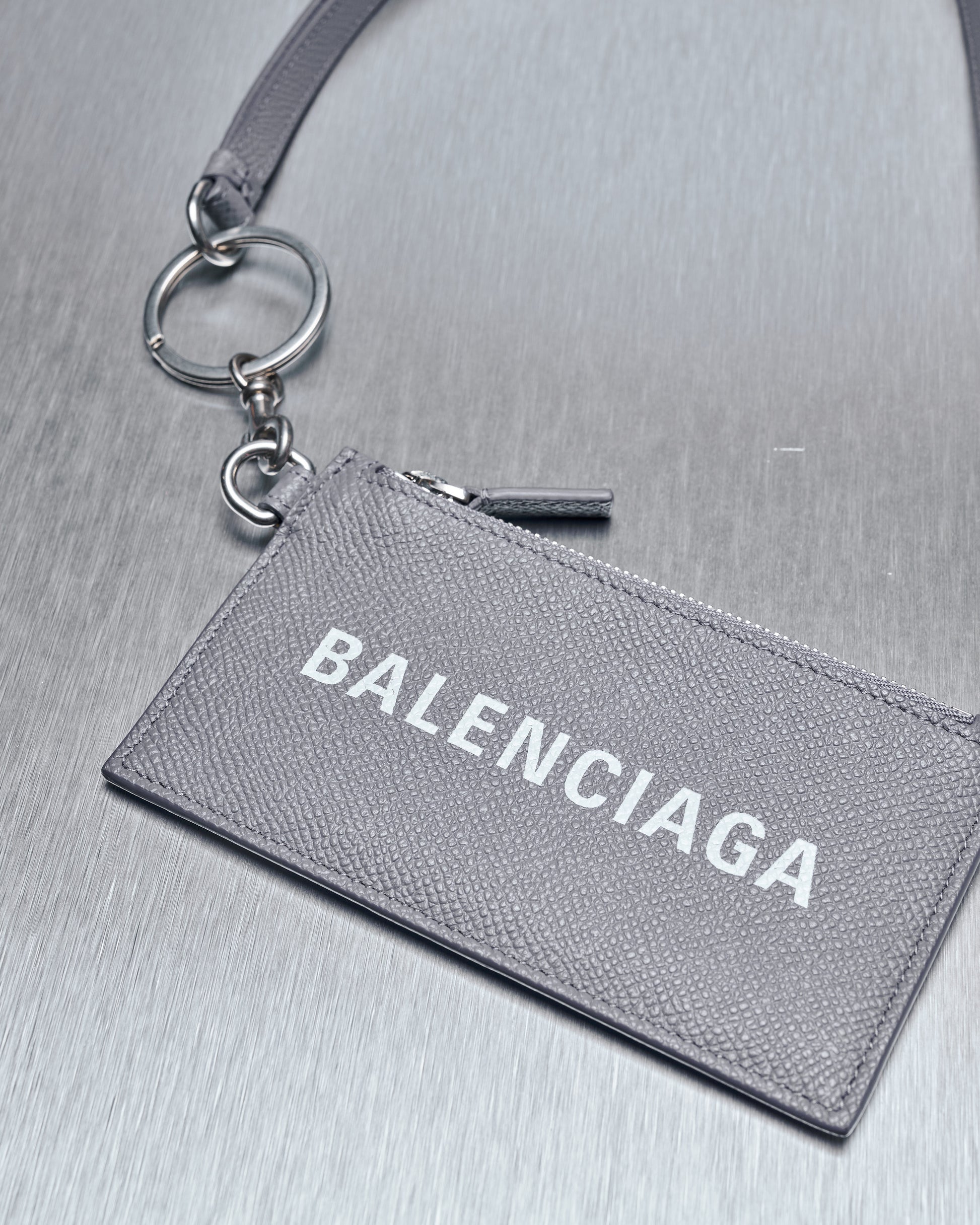 A grained leather card holder with a Balenciaga logo on the front, featuring a zip coin purse, four card slots, and a clip lanyard with a keychain.