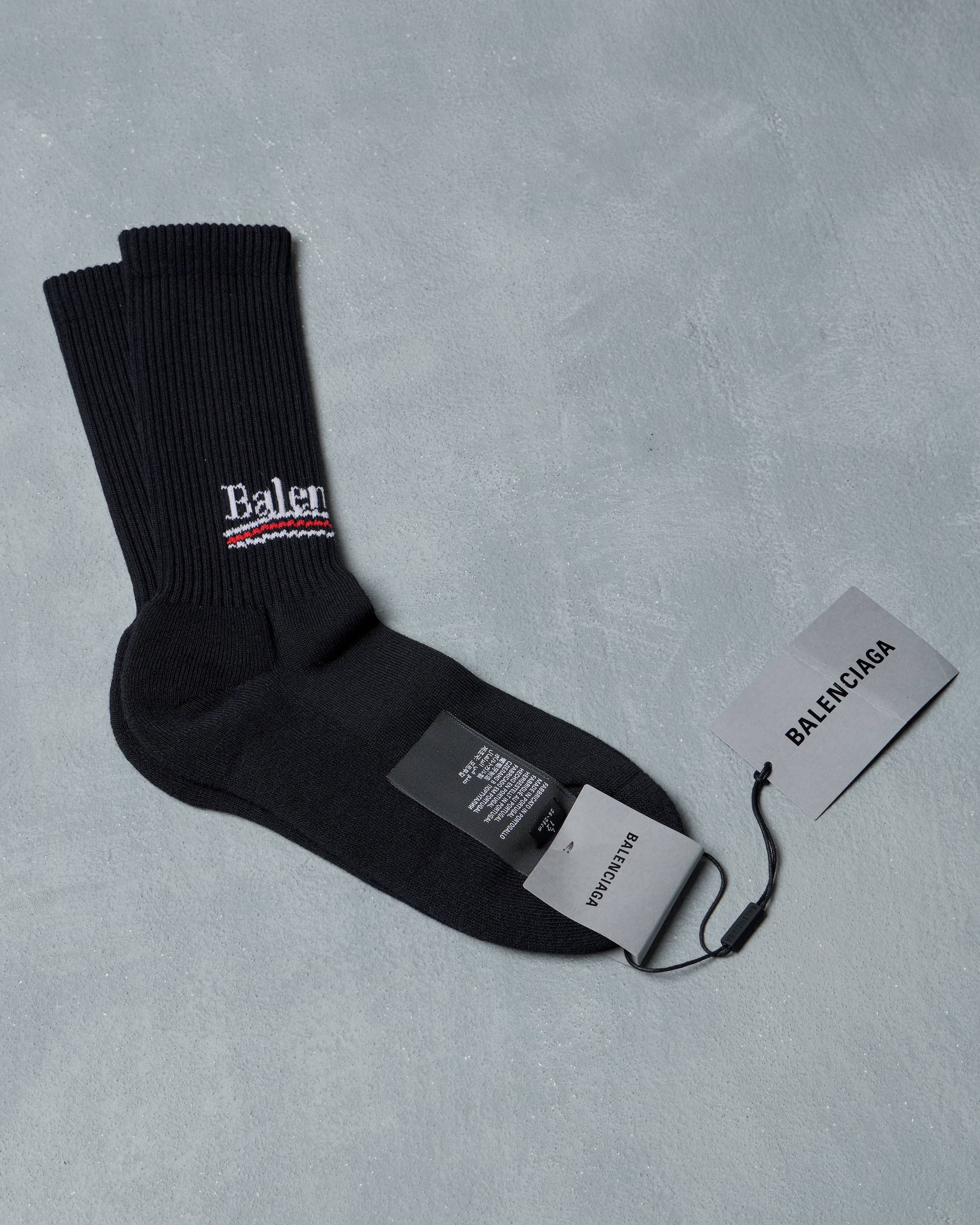 Black socks with white "Balenciaga" logo and red stripe, placed on a light gray surface. Attached tags read "BALENCIAGA," conveying luxury fashion.