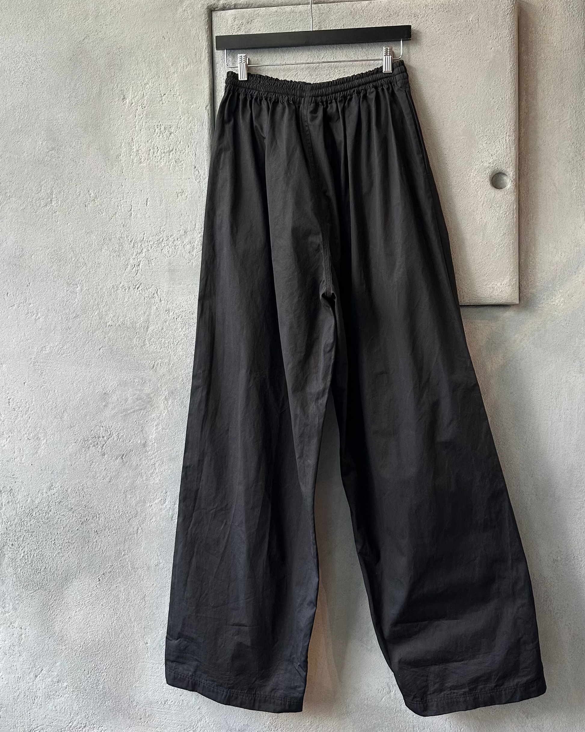 Black wide-leg Balenciaga pants hanging on a rack against a gray concrete wall.