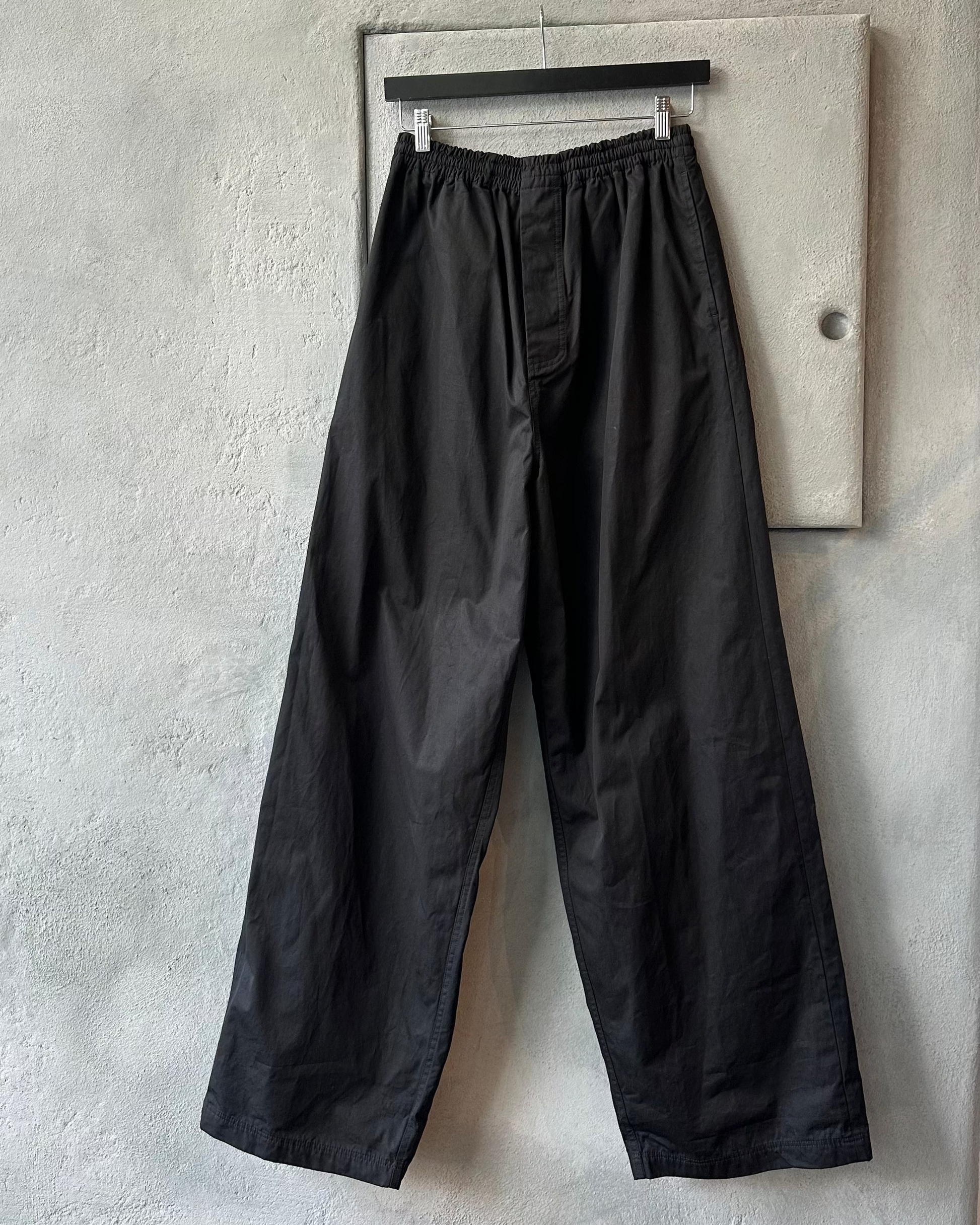 Black Balenciaga pants hanging on a wooden hanger against a gray concrete wall.