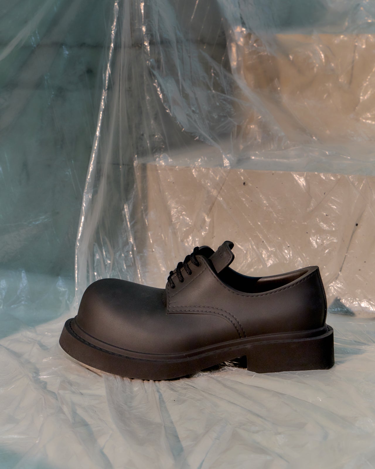 Black leather Balenciaga shoe on a light blue surface with a transparent plastic sheet in the background