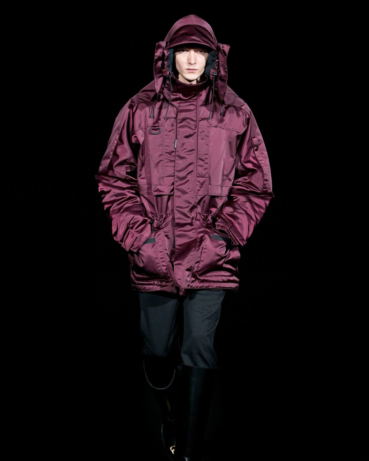 A model in a deep burgundy hooded Balenciaga parka walks confidently against a black background, exuding a sleek and modern winter style.