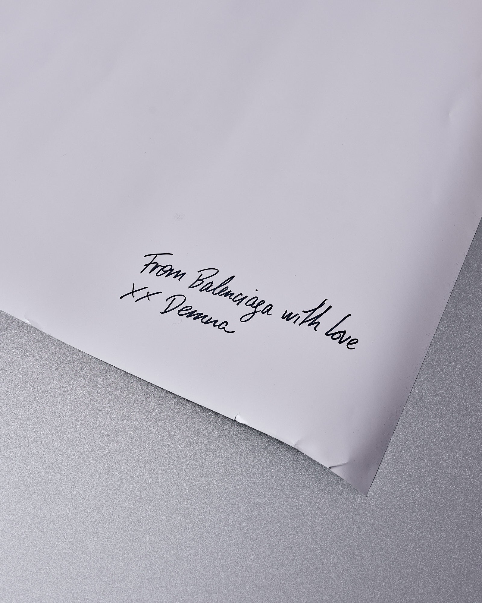 A white sheet of paper on a gray background features elegant handwriting. The message reads, "From Balenciaga with love XX Denma," conveying a personal and sophisticated tone.