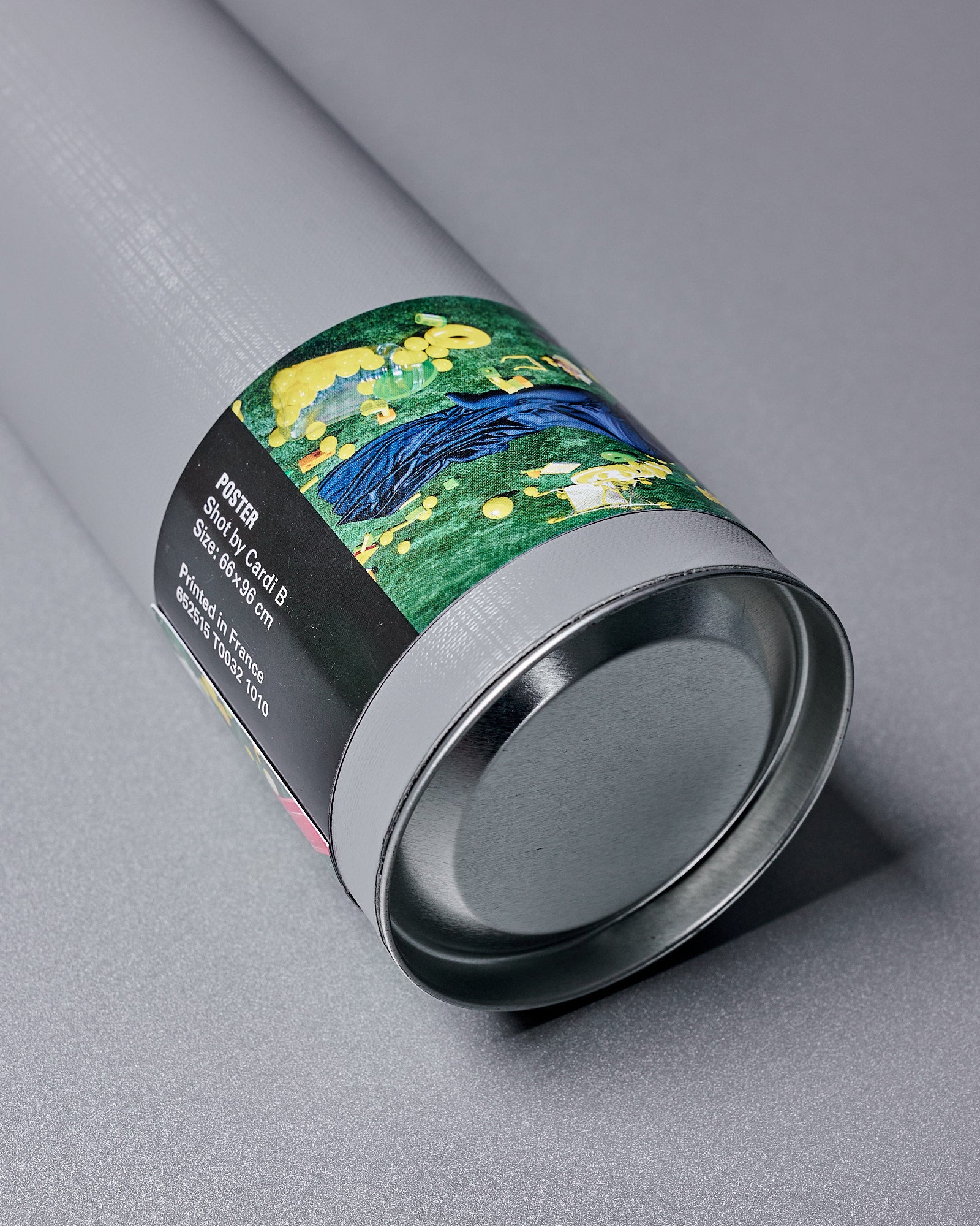 A grey cylindrical poster tube from Balenciaga x Cardi B collaboration lies on a flat surface, partially wrapped in a colourful label featuring abstract art with green, yellow, and blue tones. The label includes text details.