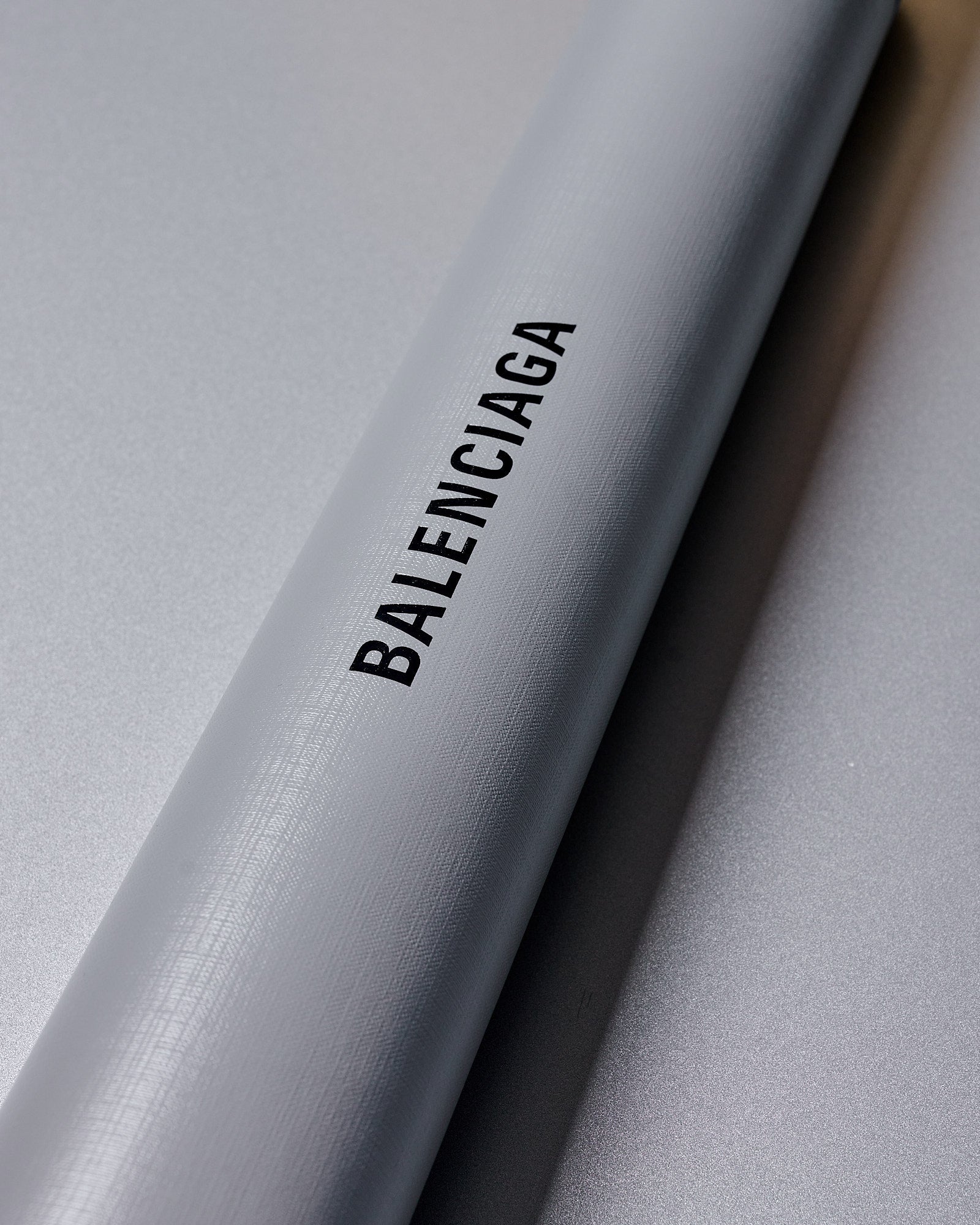 Grey textured cylindrical object with the word "BALENCIAGA" in black, angled against a smooth gray background, conveying elegance and luxury.