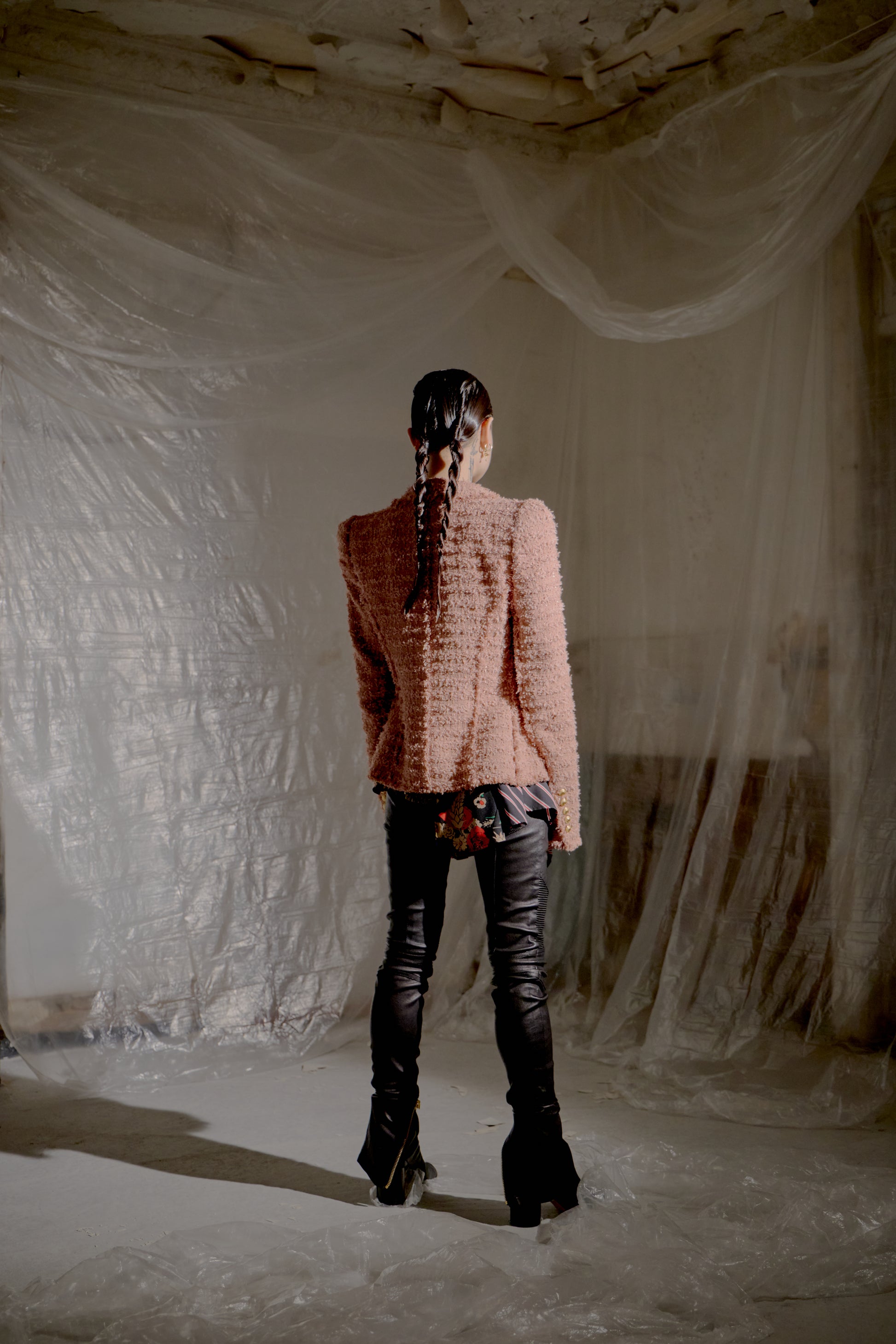 Person wearing a pink Balmain jacket and black pants standing in a room with white plastic sheeting.