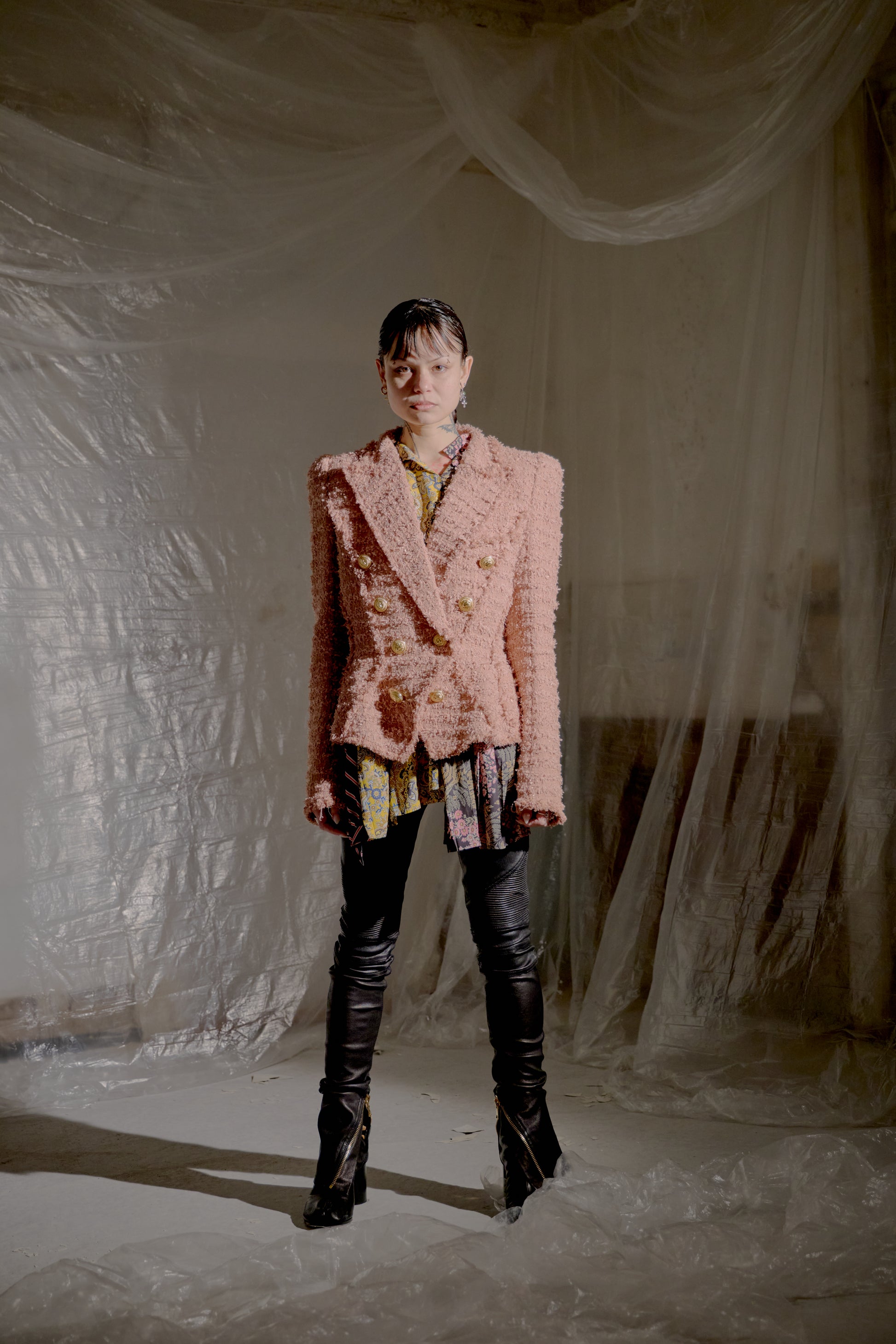 Person wearing a pink textured Balmain jacket and black boots in a draped fabric setting