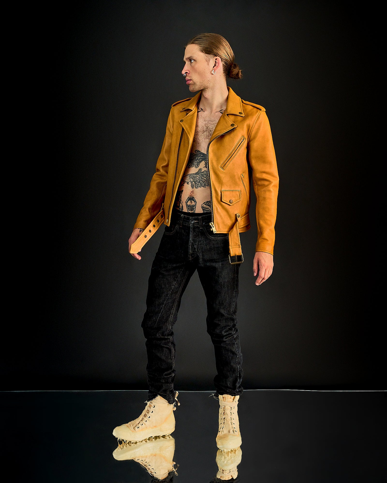 A person stands against a dark background, wearing a tan leather Blackmeans jacket, black jeans, and beige boots. The jacket is open, revealing tattoos. The tone is edgy.