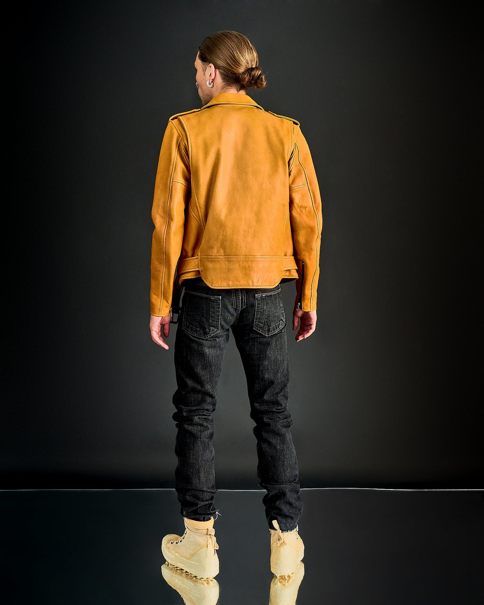 Person with tied back hair wearing a mustard leather Blackmeans jacket, black jeans, and beige boots stands facing away against a dark backdrop.