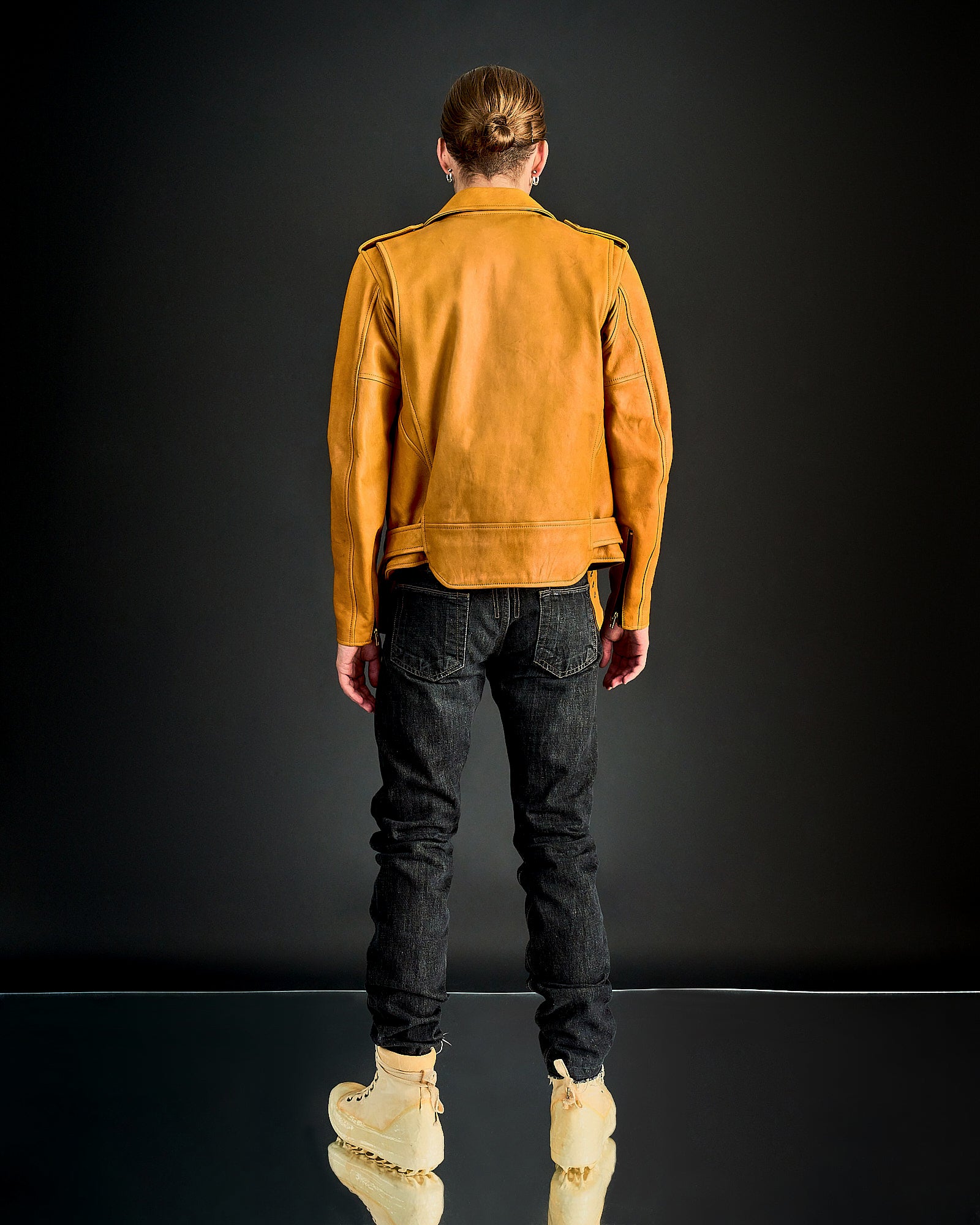 Person with tied back hair wearing a mustard leather Blackmeans jacket, black jeans, and beige boots stands facing away against a dark backdrop.