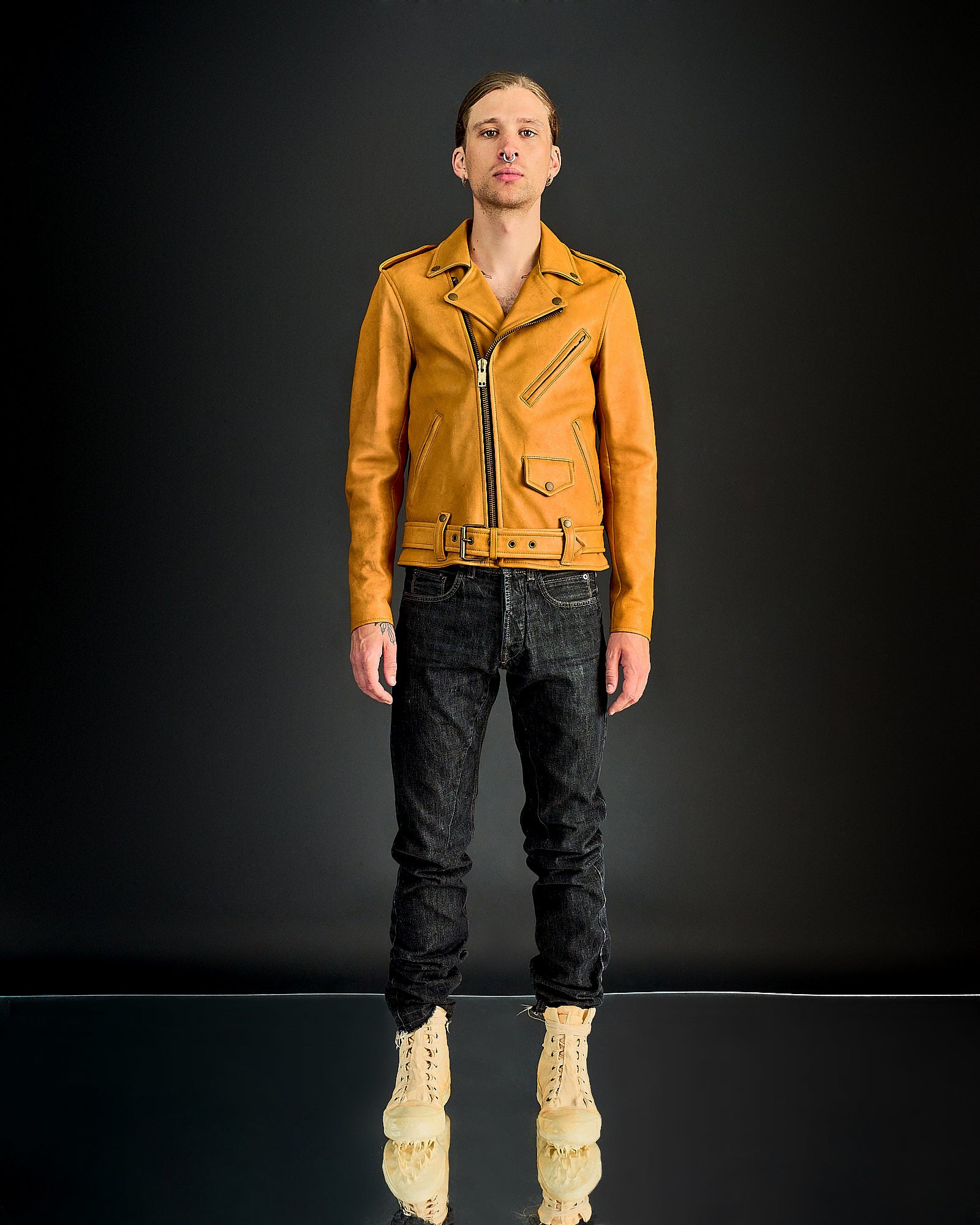 A person stands confidently against a dark backdrop wearing a mustard leather Blackmeans jacket, dark jeans, and beige lace-up boots, projecting a trendy, edgy style.