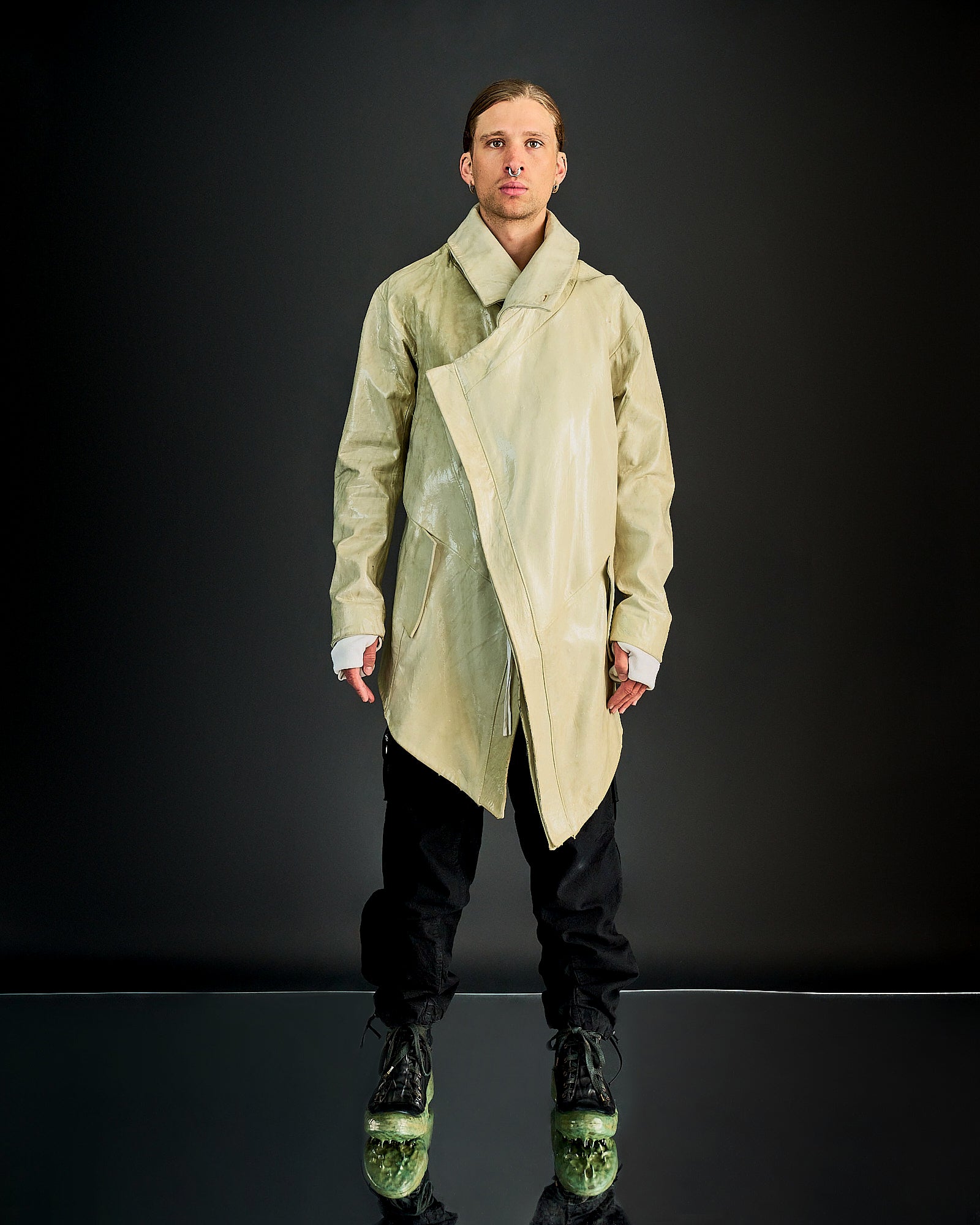 Person in a light beige, Boris Bidjan Saberi asymmetrical coat stands against a dark backdrop. They're wearing black pants and distinctive green-soled boots.