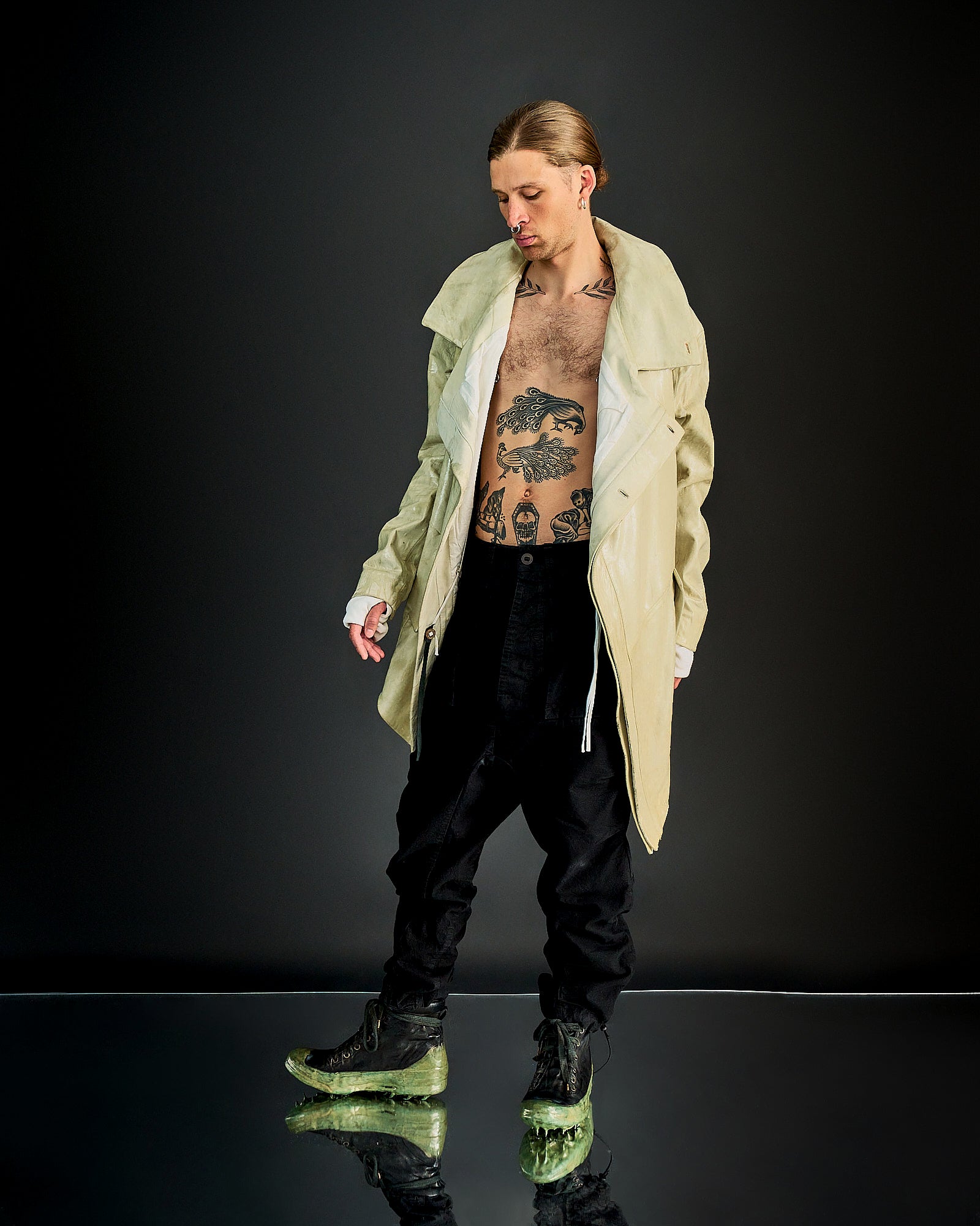 A person with a contemplative expression wears an open beige Boris Bidjan Saberi coat revealing tattoos on their torso, black pants, and green-soled shoes against a dark background.