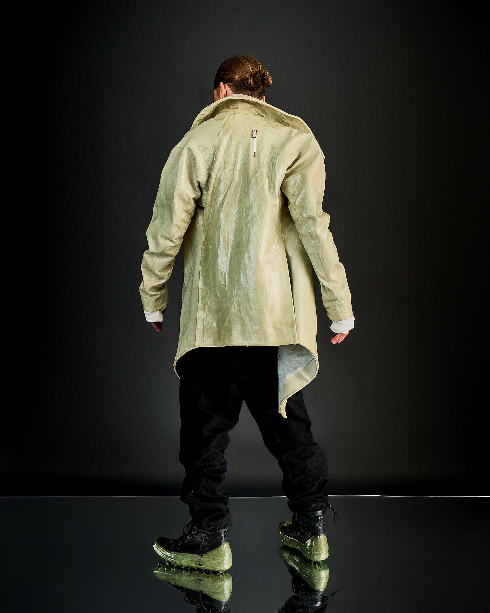 Person in a light Boris Bidjan Saberi coat faces away, wearing black pants and green transparent-sole shoes. The setting is a reflective, dark-toned surface.